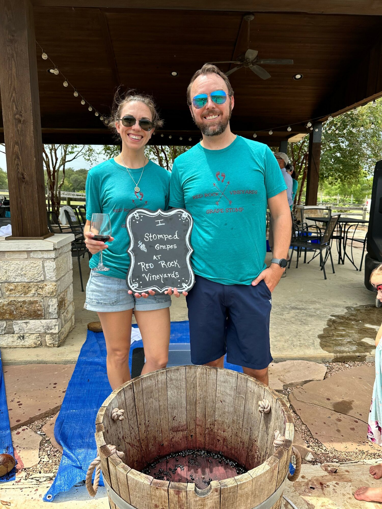 Two women stand outdoors near a fire pit, holding a chalkboard sign, smiling, with trees and outdoor furniture in background.