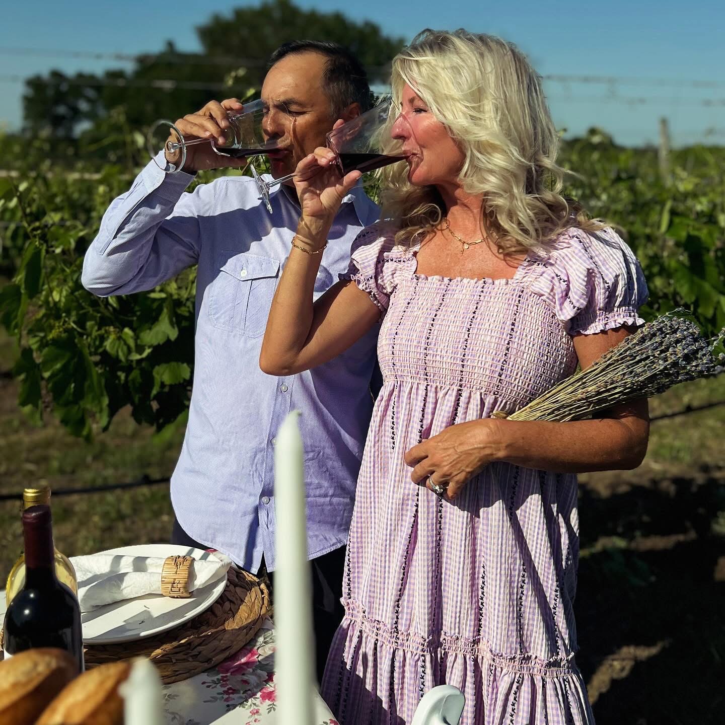 Two people drinking wine outdoors, one woman holding a bundle of sticks, vineyard background, sunny day.