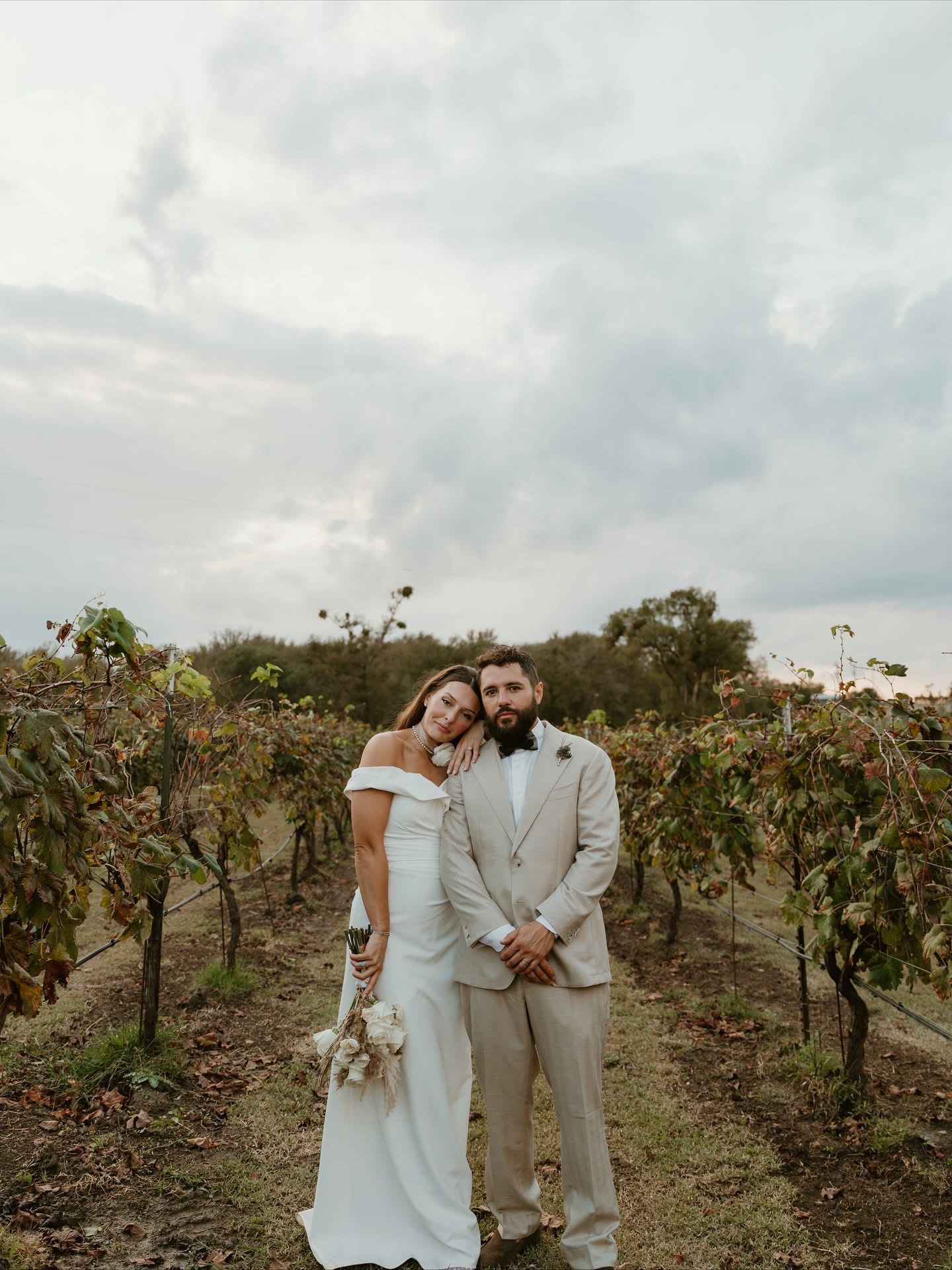 A couple in wedding attire standing in a vineyard with cloudy sky above, holding flowers, facing camera.