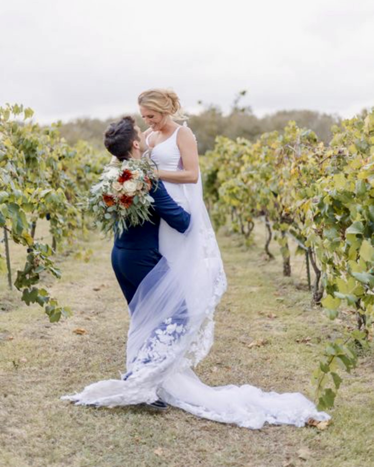 A bride and groom in a vineyard, with the groom holding a bouquet, standing close and looking at each other.