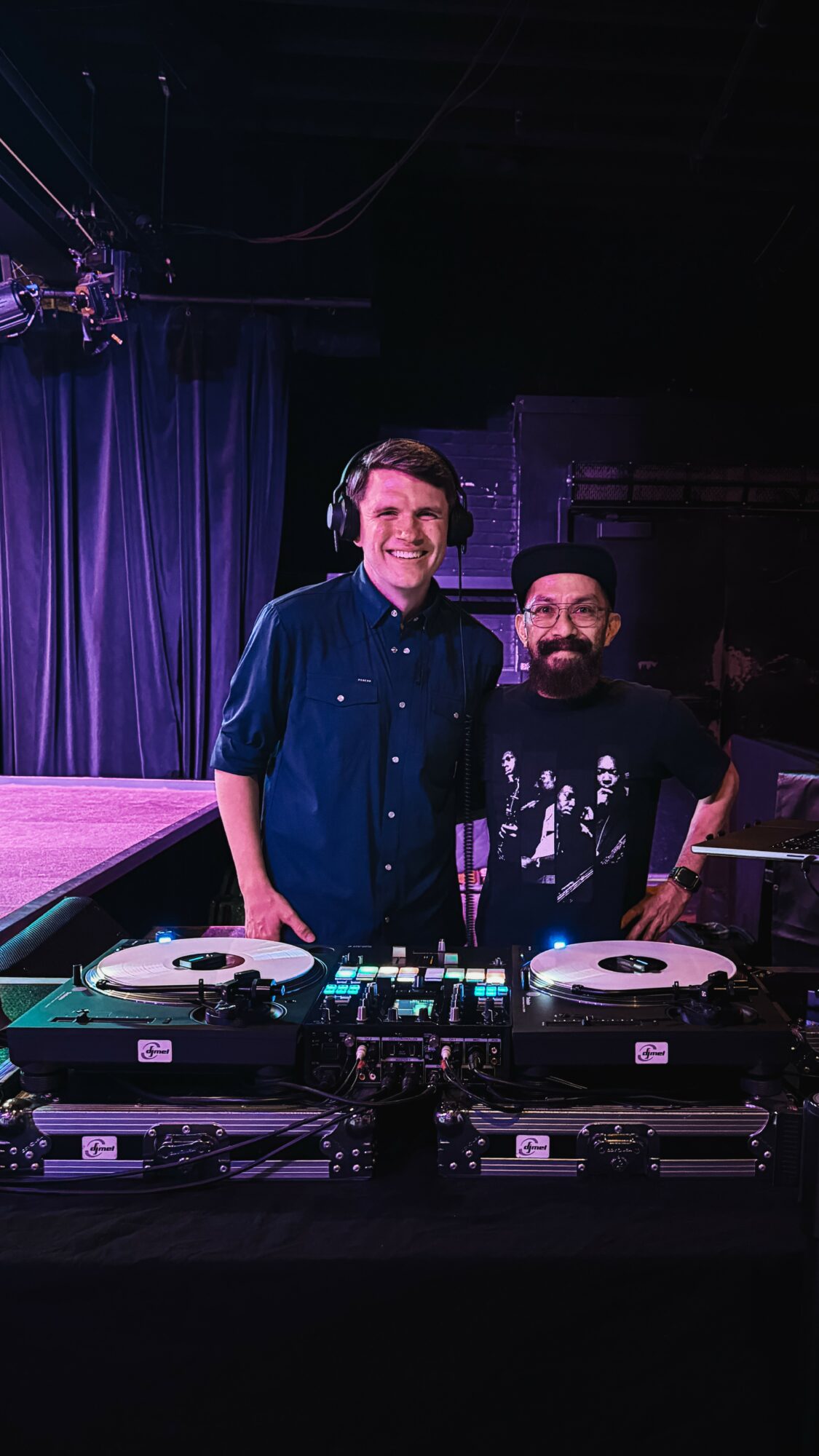 Two men standing behind DJ equipment on stage, smiling, with dark curtains in the background.