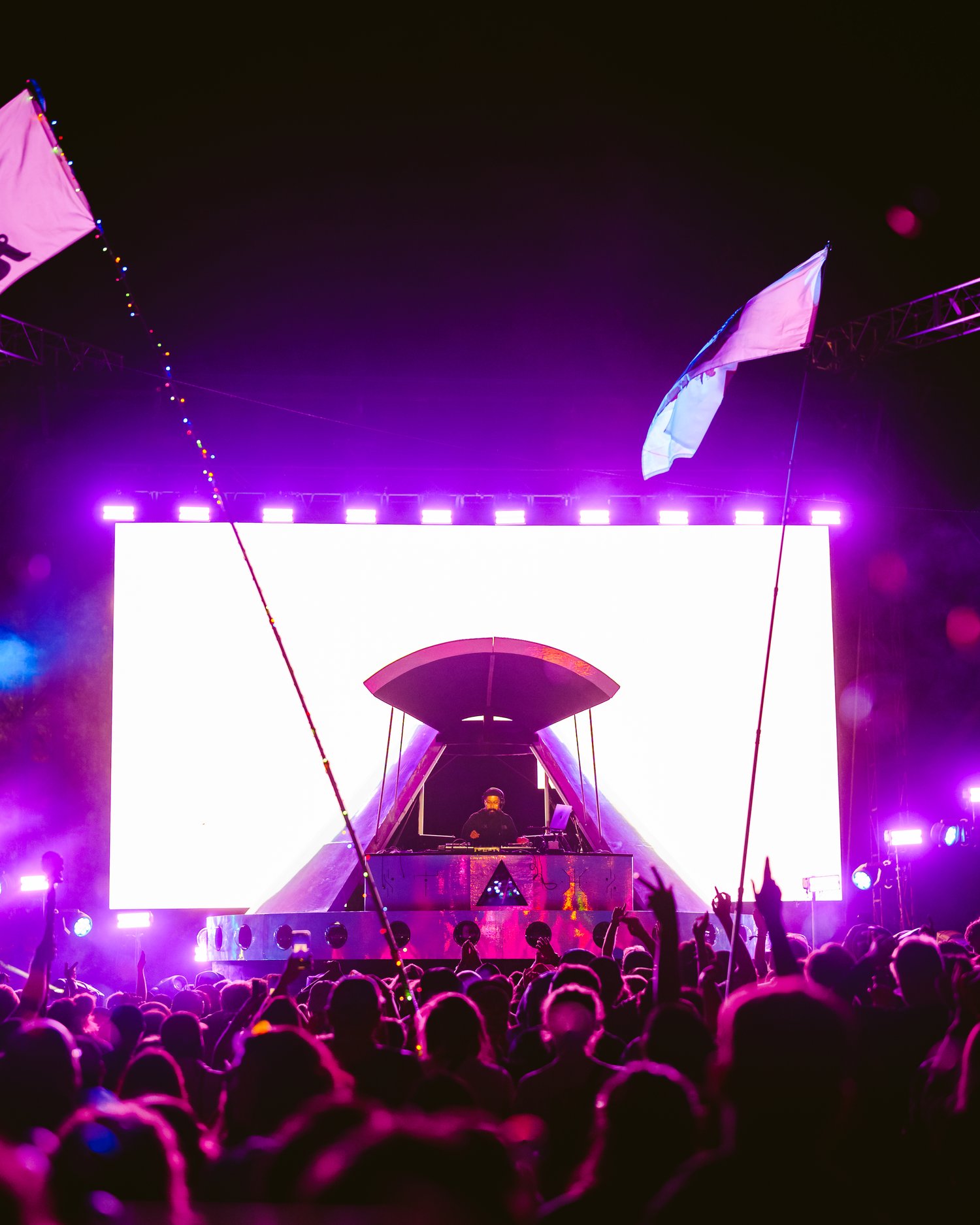 Crowd watching a large illuminated stage with purple and white lights at night, flags waving above the audience.