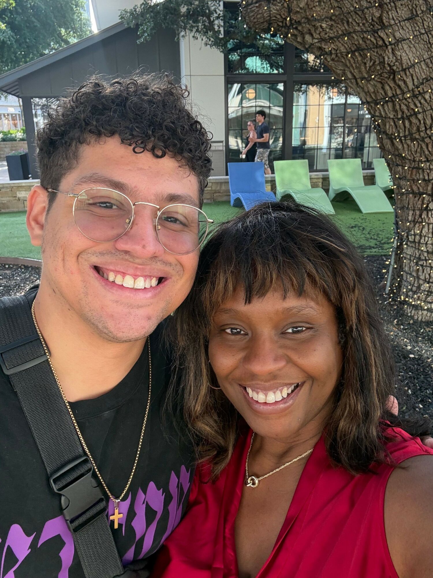 Two smiling people, one with glasses and curly hair, and the other with straight hair, outdoors near a tree and building.