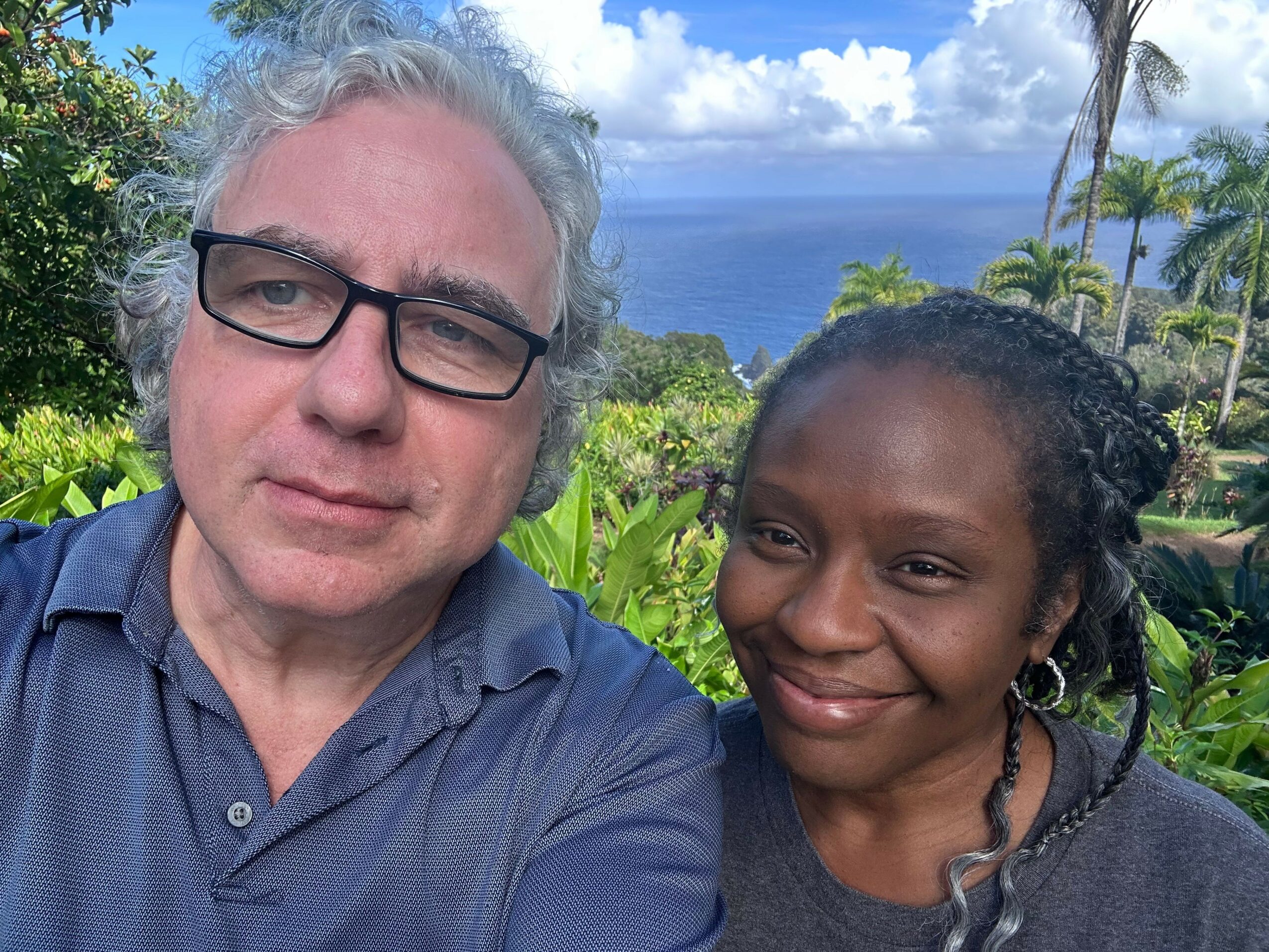 Two people outdoors, one with glasses and gray hair, the other with dark hair, smiling with greenery and sky in background.