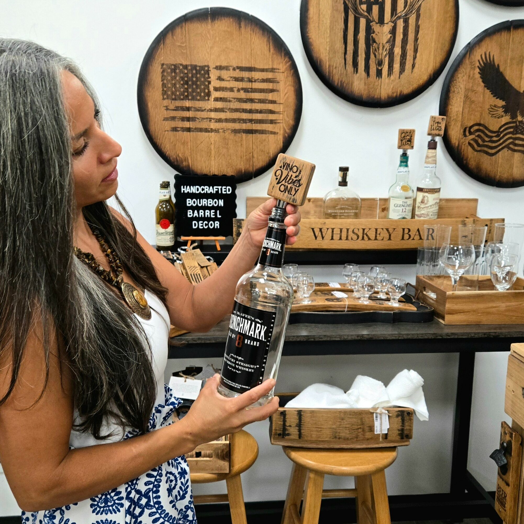 Woman with long gray hair holding a bottle of alcohol in a store with whiskey-themed wall art and bar accessories.