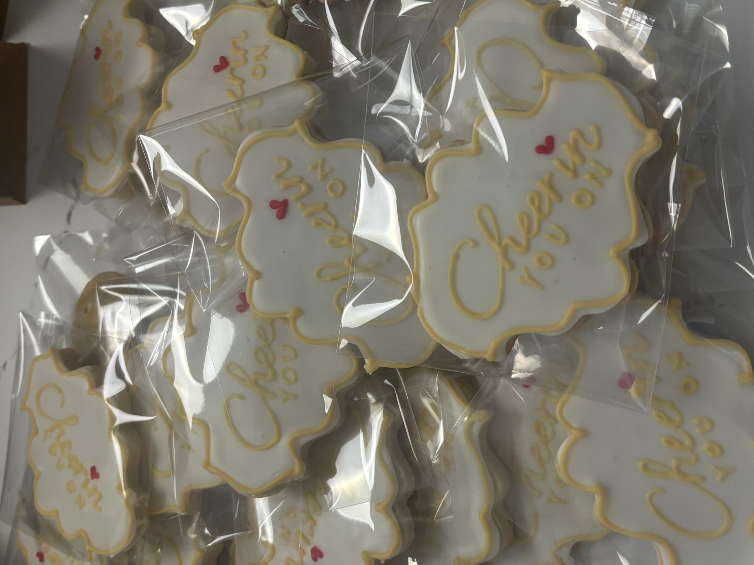 Cookies decorated with white icing and gold writing, packaged in clear plastic bags, with small red hearts, on a dark surface.