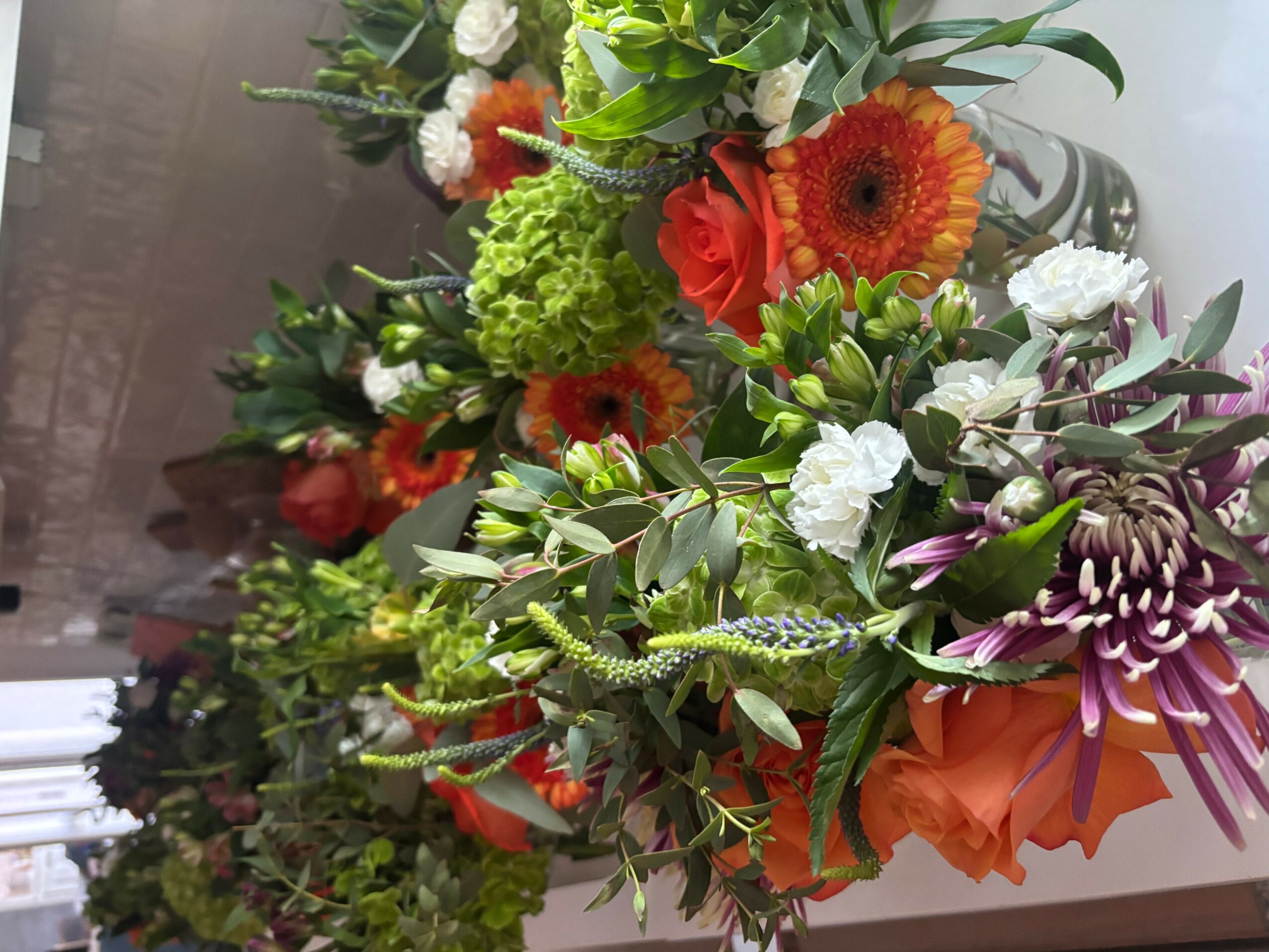 Colorful bouquet of flowers with orange, white, purple, and green blooms and foliage, arranged on a white surface.