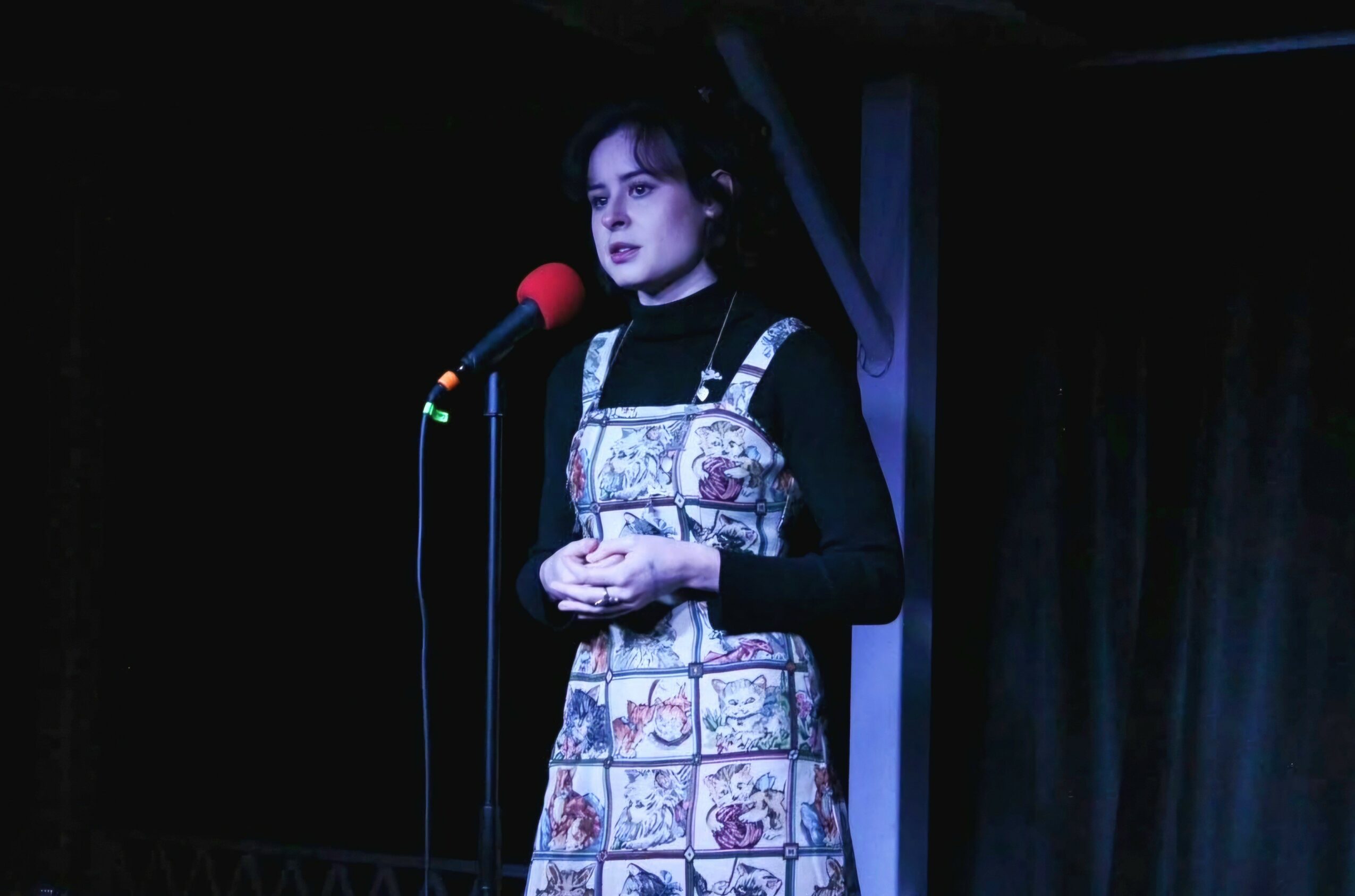Woman standing on stage with a microphone, wearing a patterned apron over a black top, in a dark setting.