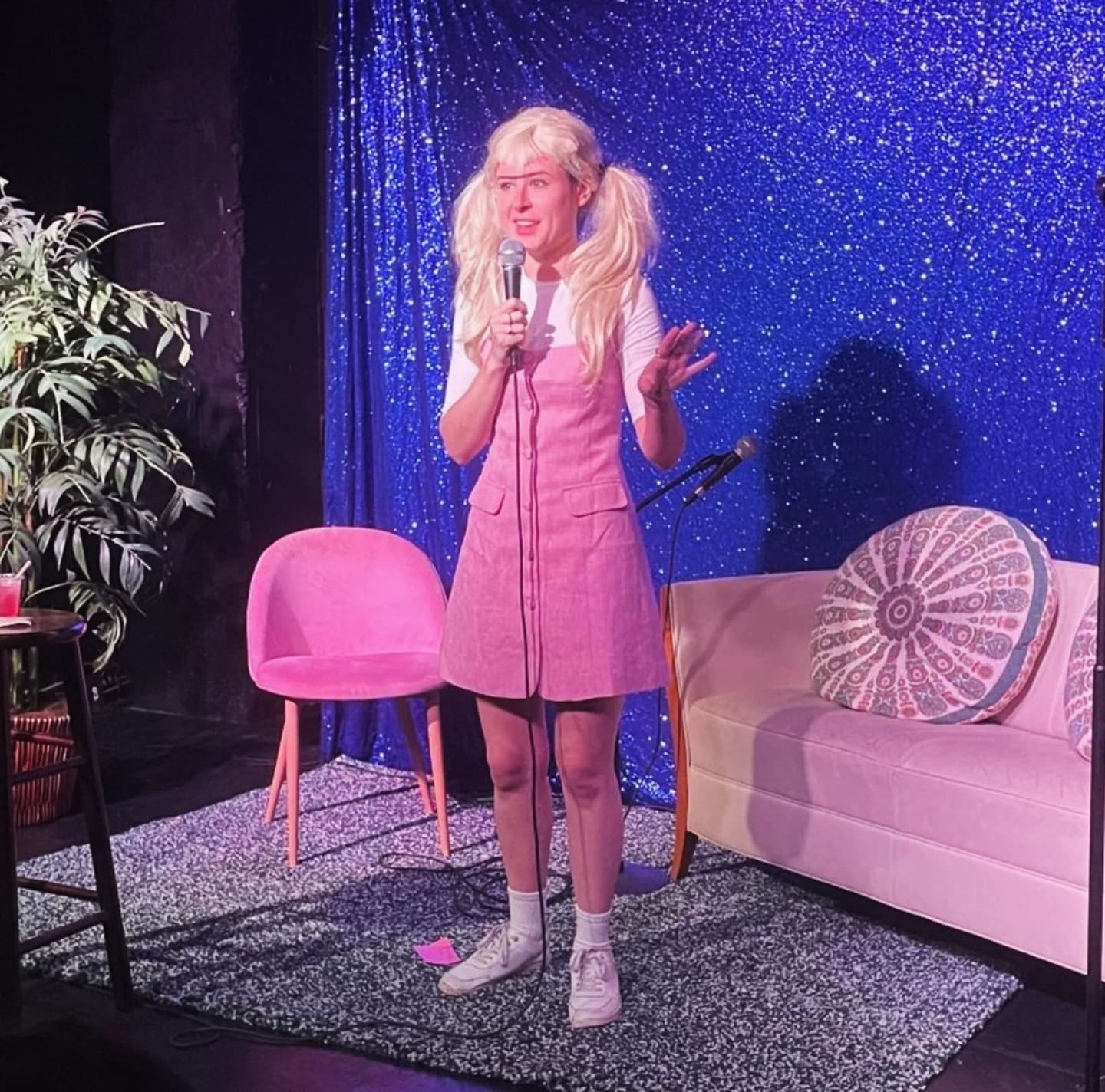 Person with long blonde hair in pigtails, pink dress, holding microphone, standing on stage with pink chair and sofa, blue sparkly backdrop.