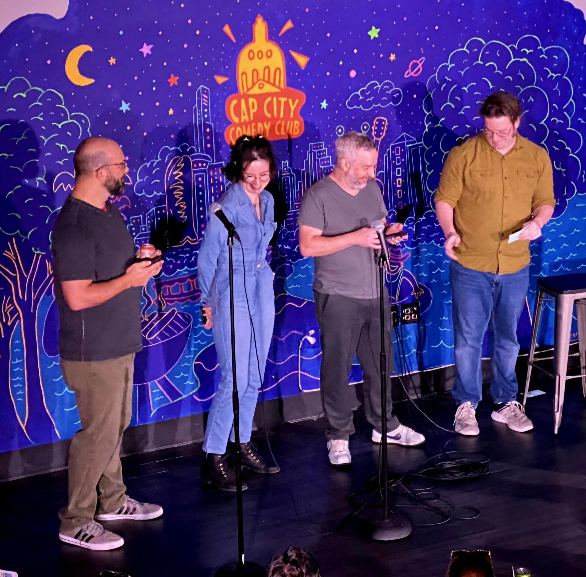 Four people stand on stage with microphones, in front of a colorful cityscape backdrop with stars and moon, performing or speaking.