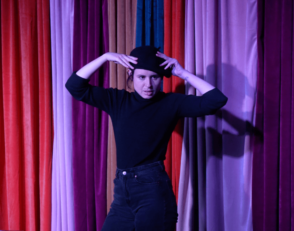 Person with hands on head standing in front of colorful curtains, wearing black clothing, looking at camera.