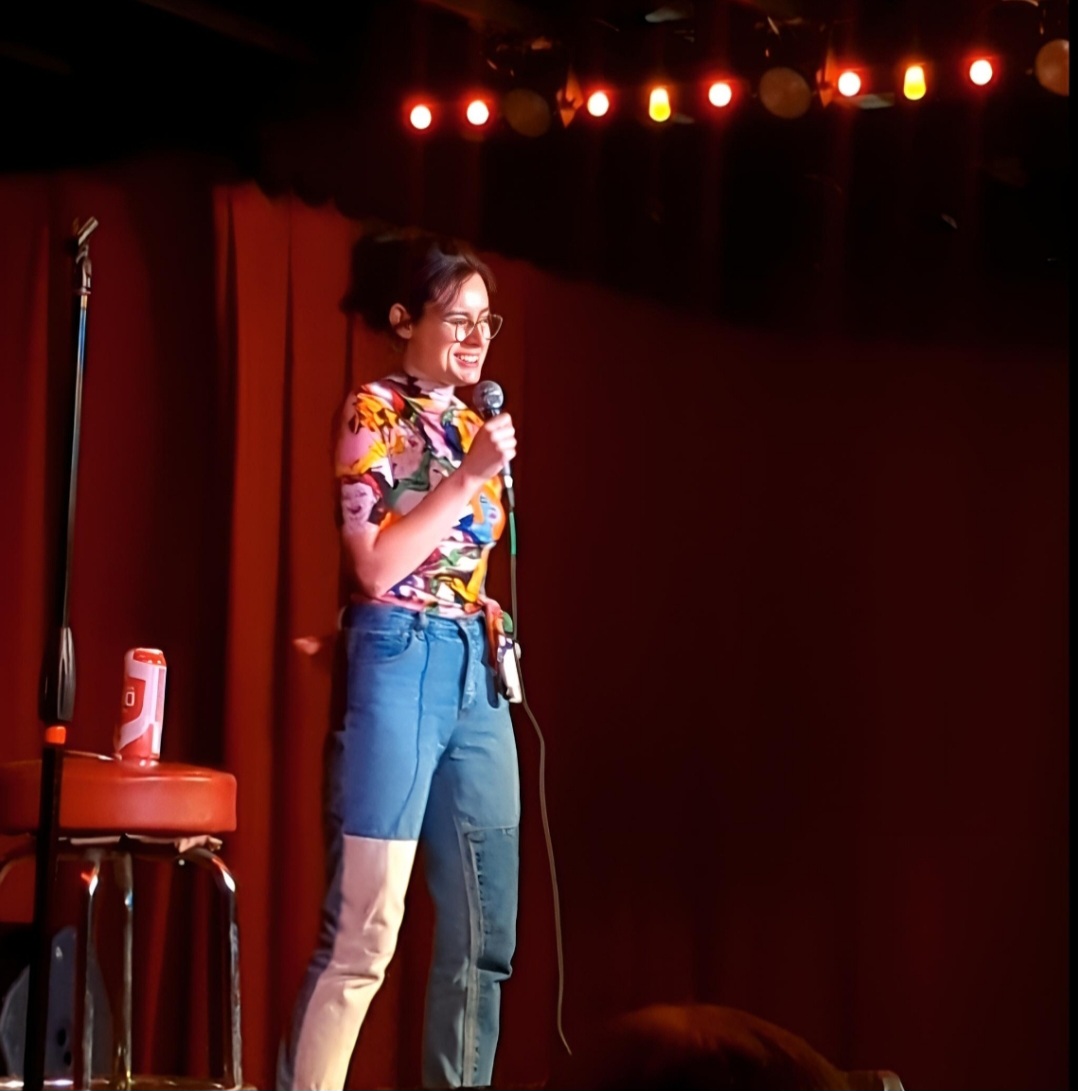 Young woman with glasses holding a microphone on stage with red curtains and stage lights behind her.