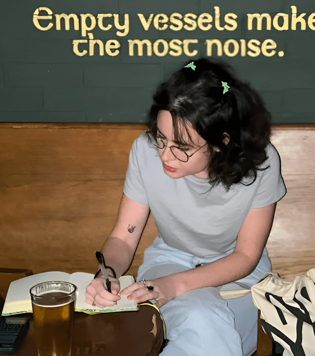 Woman with glasses writing in a notebook at a table with a glass of beer, dark green wall behind her.