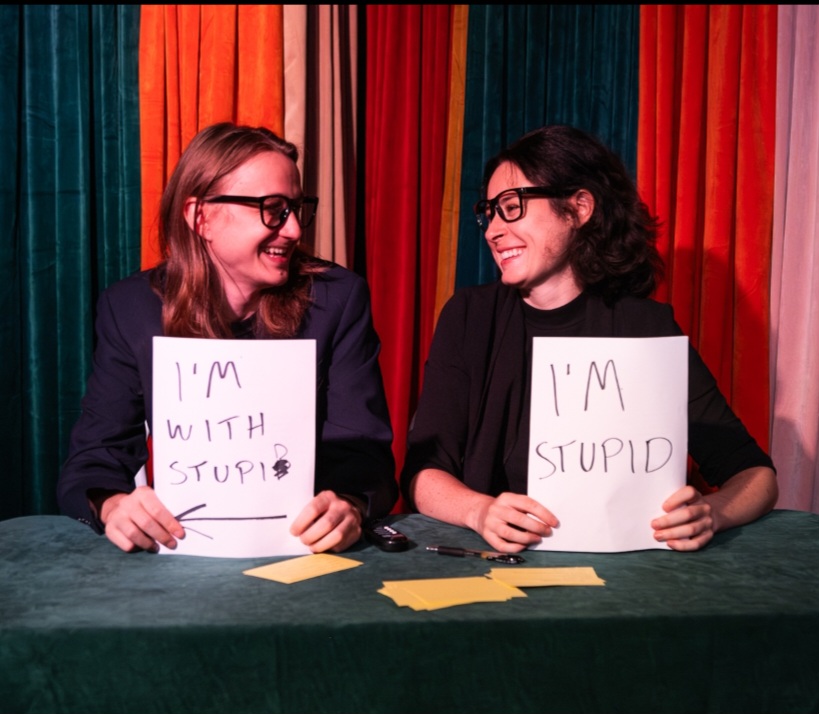 Two women sitting at a table holding signs with humorous messages, smiling at each other, colorful curtains in the background.