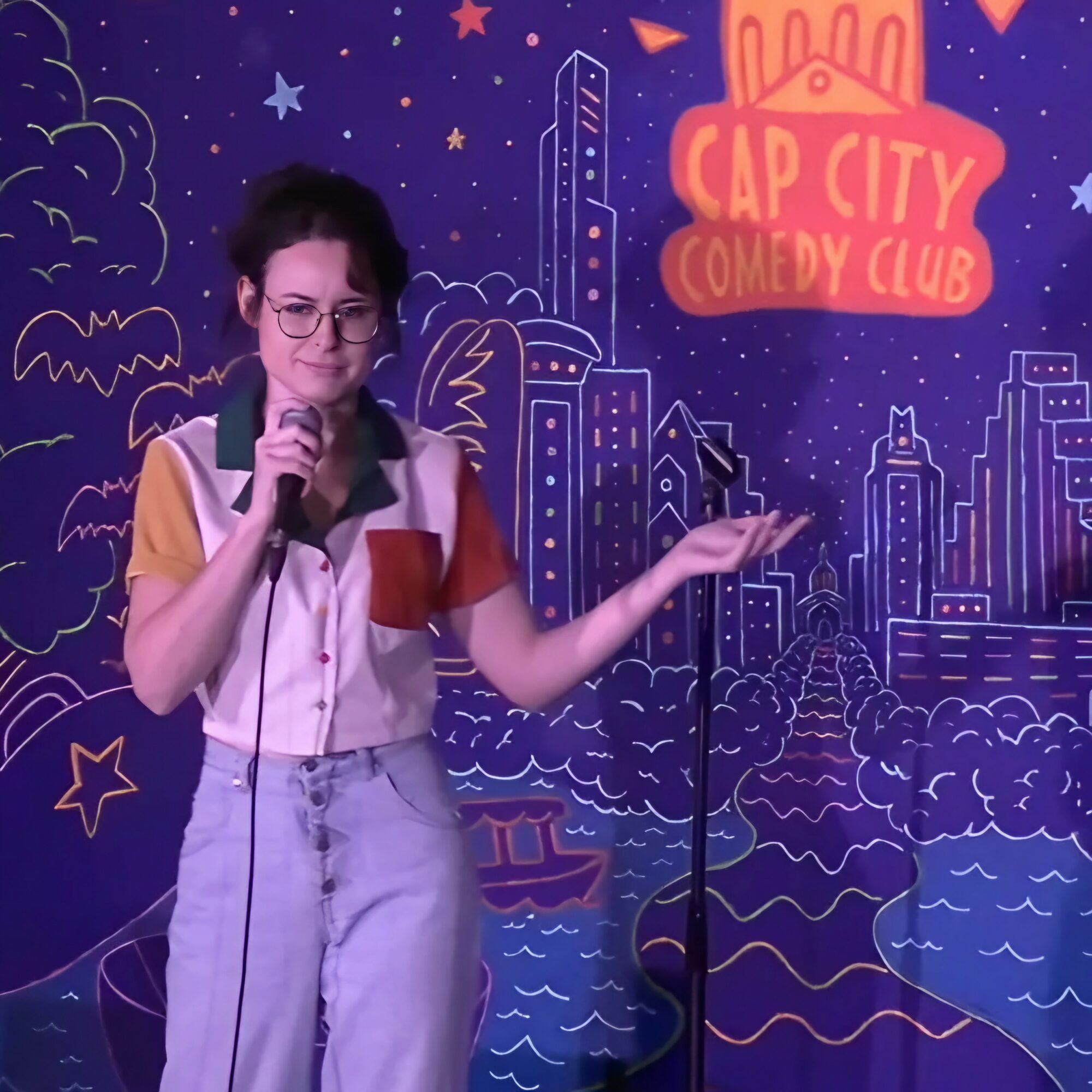 Woman with glasses holding a microphone on stage at Cap City Comedy Club, colorful cityscape background.