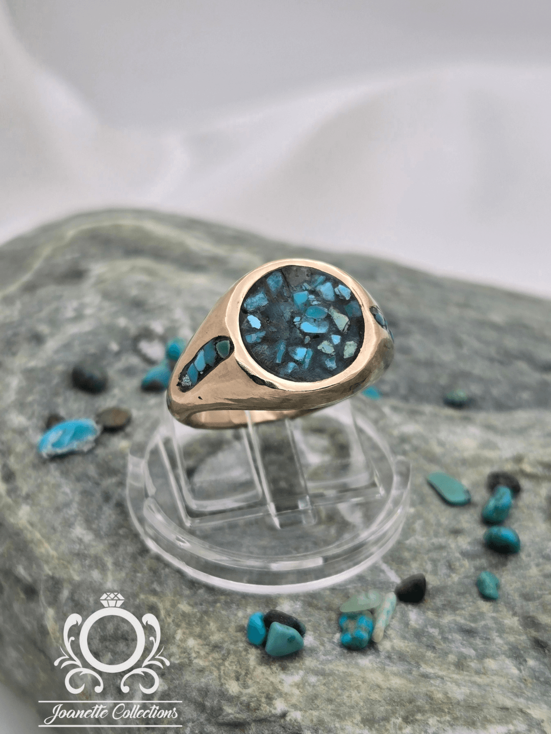 Gold ring with a large blue gemstone, displayed on a clear stand with scattered blue and black stones around, on a textured surface.