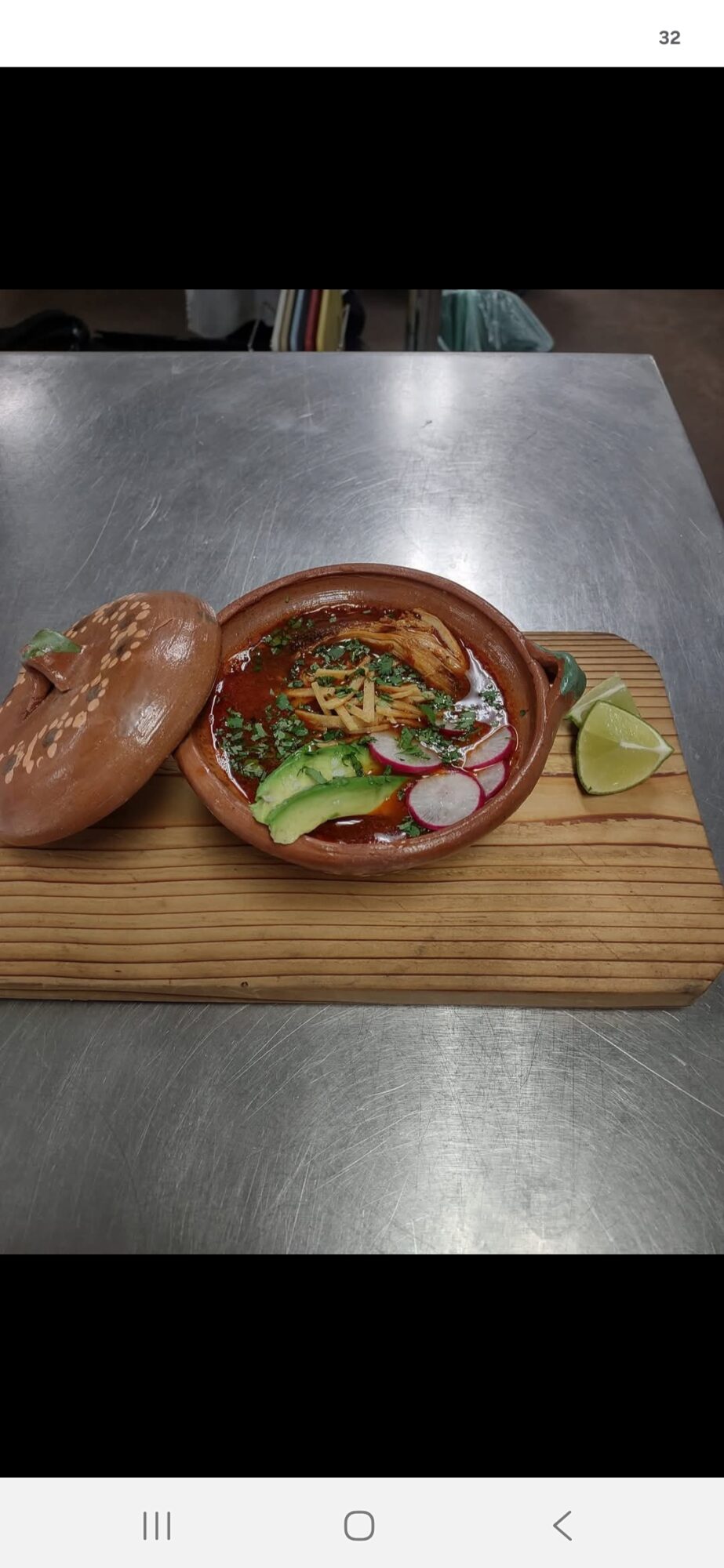 Bowl of food with vegetables and sauce on a wooden board, with a bread roll and lime wedge, on a metal surface.