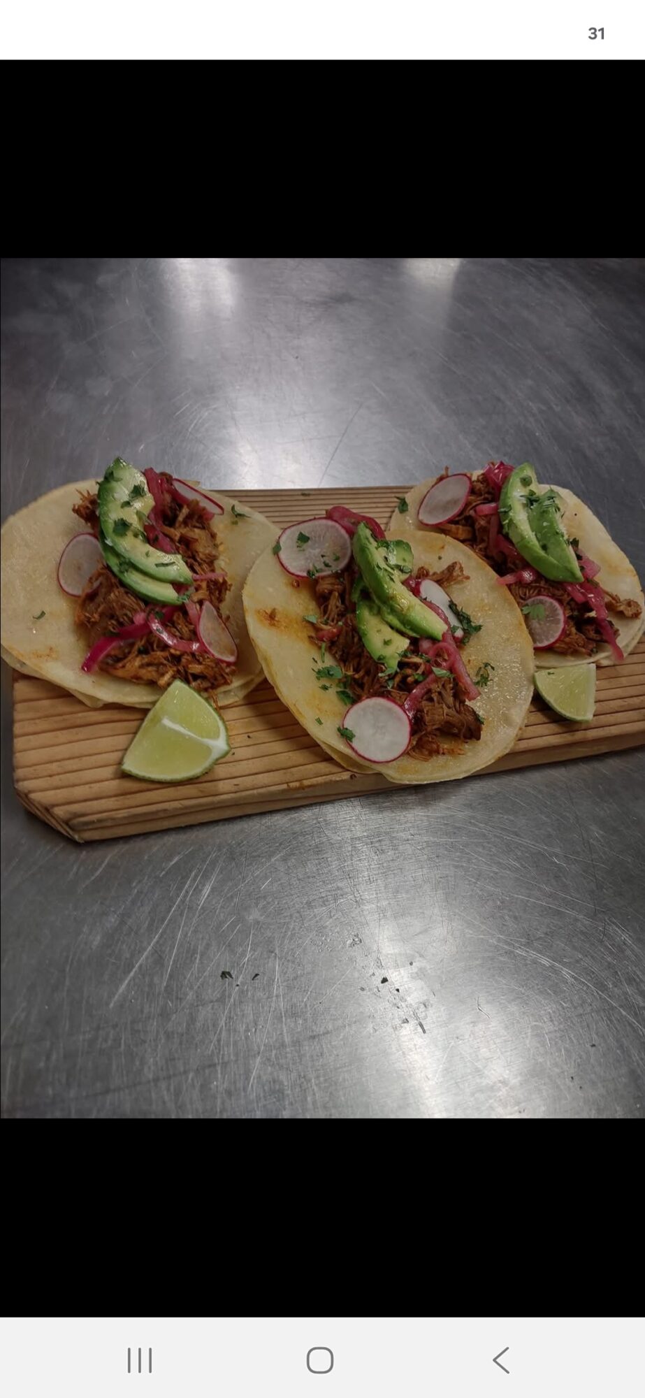 Three tacos with meat, avocado slices, radish, and lime on a wooden board, on a metallic surface.