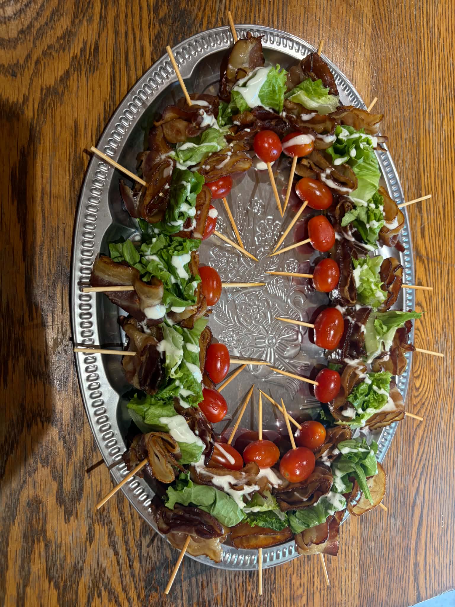Oval platter with skewers of cherry tomatoes, lettuce, and meat, garnished with greens, on a wooden table.