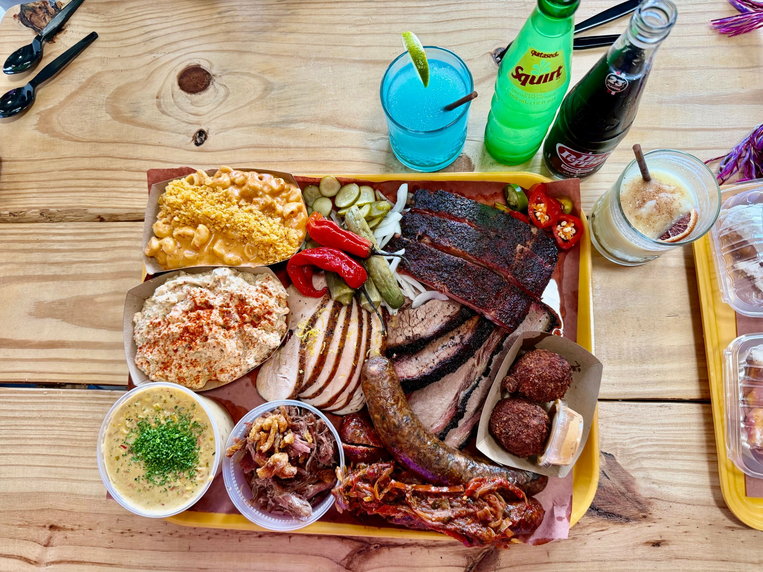 Wooden table with a tray of grilled meats, vegetables, and side dishes, along with drinks and condiments.