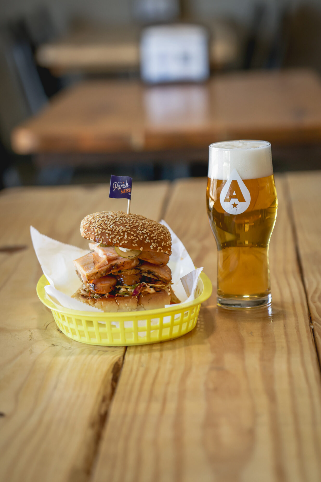 Sandwich with sesame bun, lettuce, and meat in a yellow basket, next to a glass of beer on a wooden table.