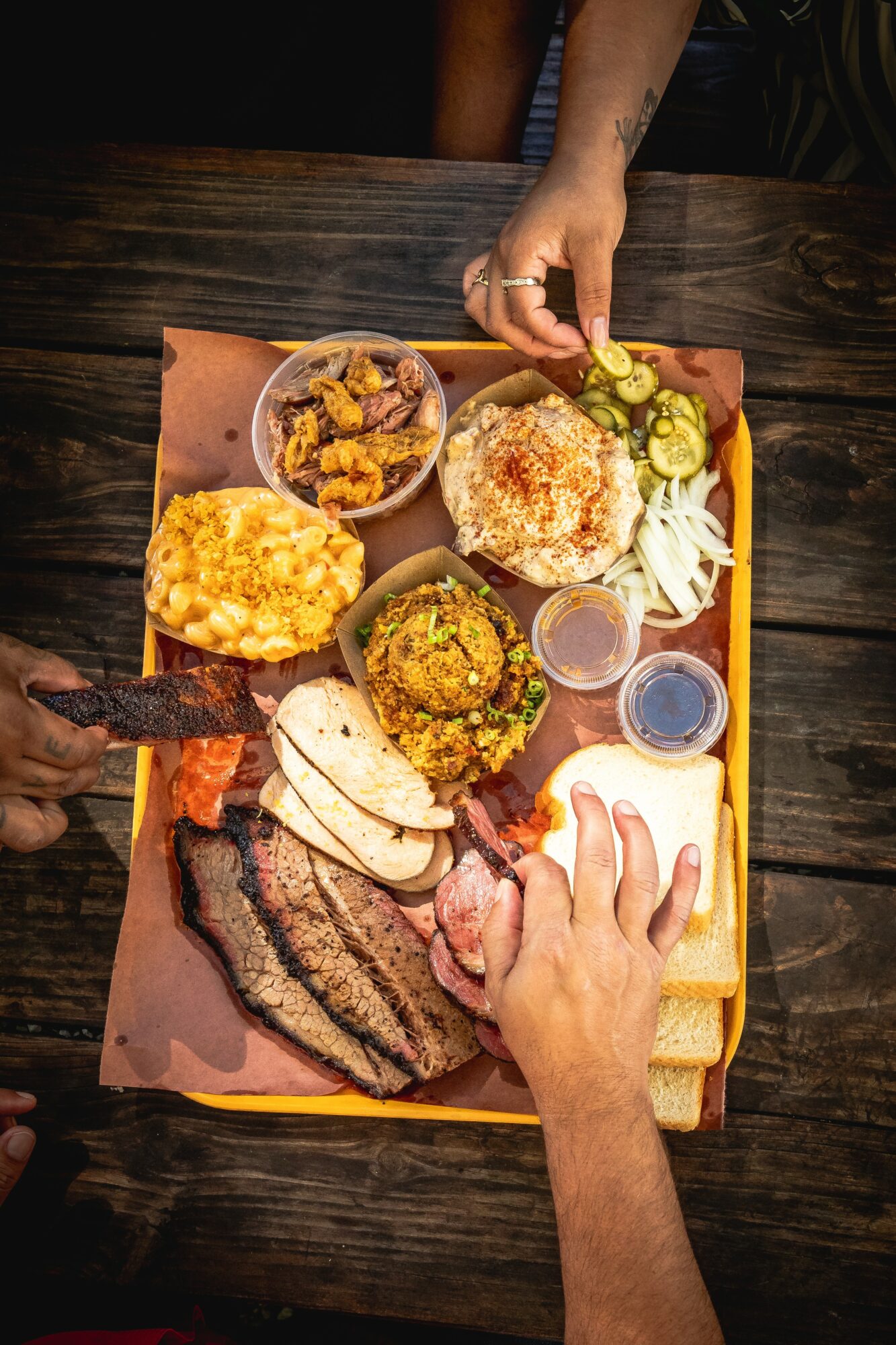 Tray with rice, grilled meats, vegetables, and side dishes on a dark wooden table, hands reaching for food.
