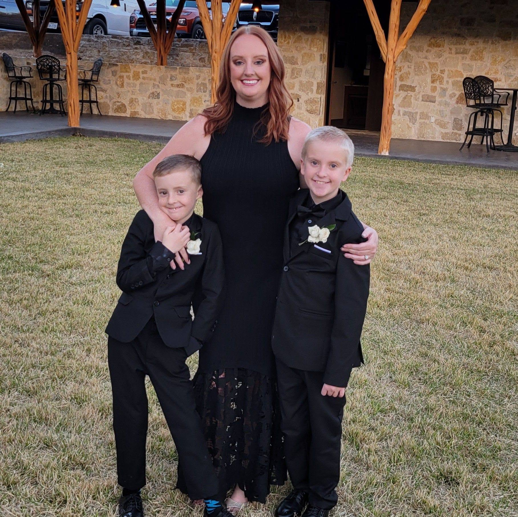 Woman with two boys in formal attire standing outdoors on grass, smiling, with a stone building in background.