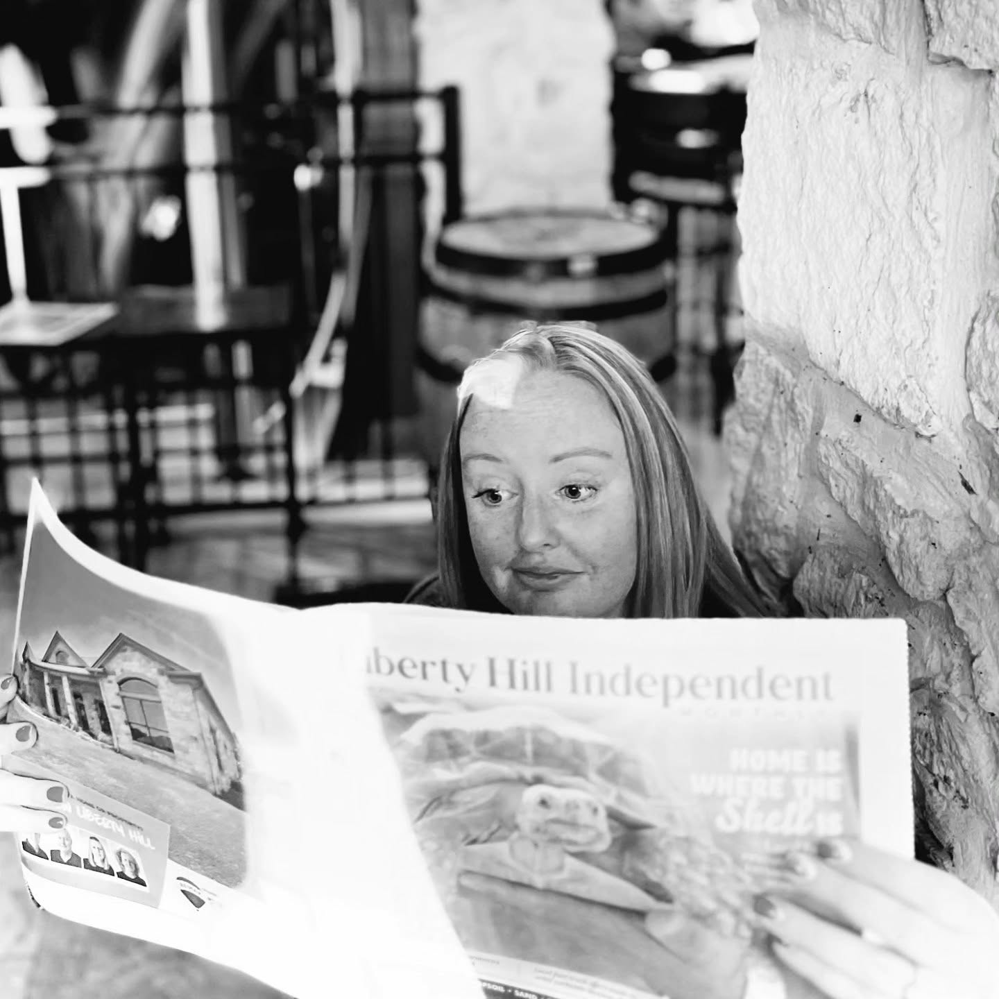 Woman reading a newspaper outdoors near a stone wall, with chairs and tables in the background.