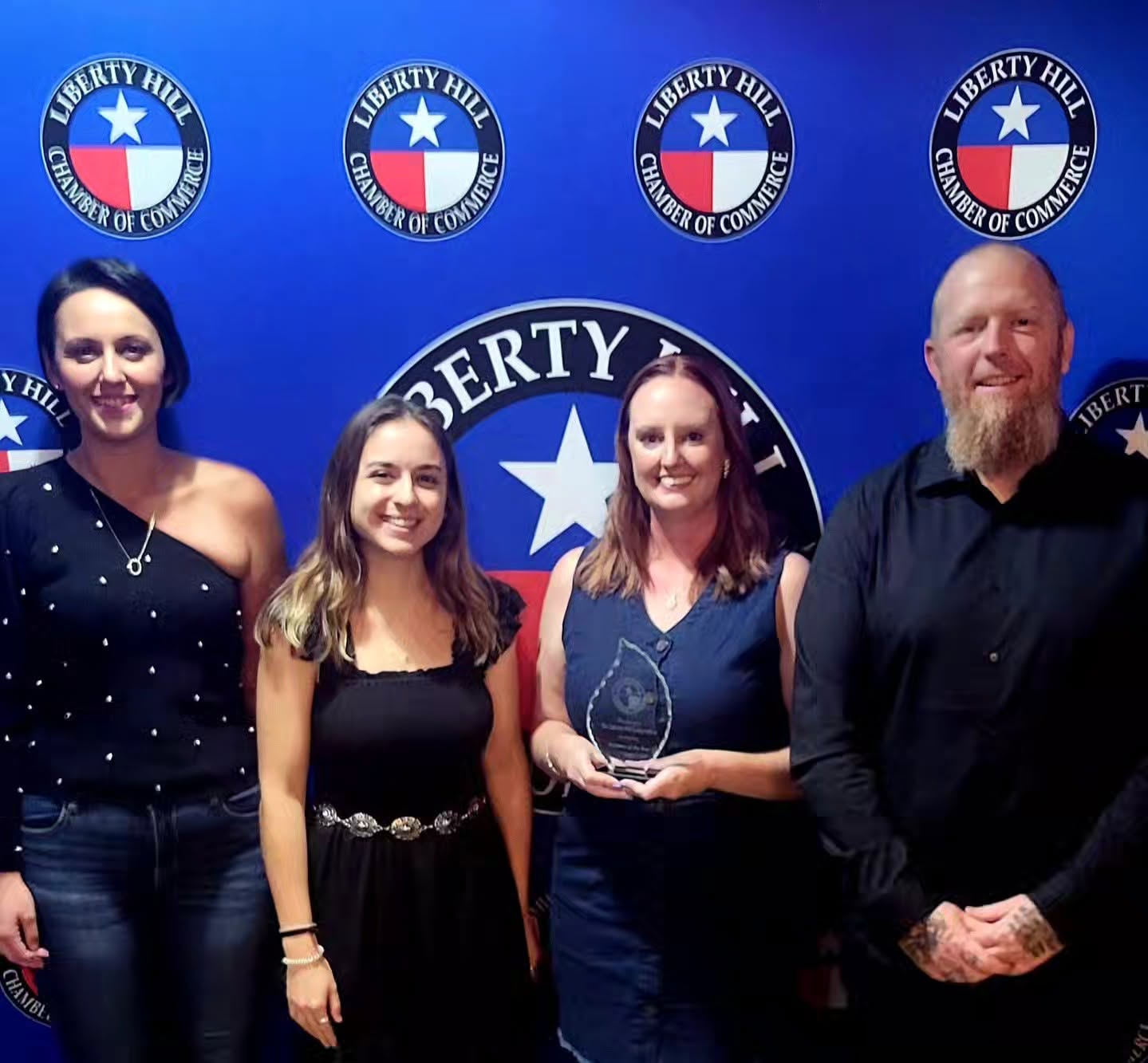 Four people standing in front of a blue background with Liberty Hill logos, one holding an award, smiling.