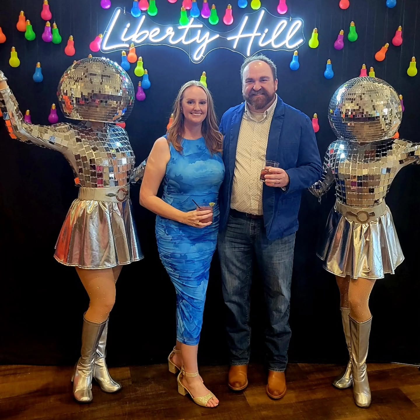 Two people stand between two performers in shiny costumes with mirrored helmets, in front of a colorful backdrop with 'Liberty Hill' sign.