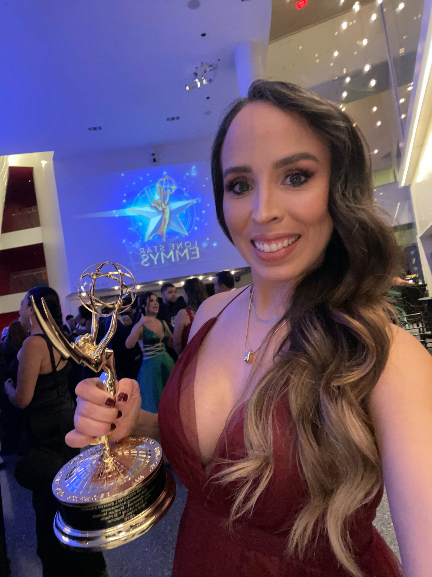 Woman with long wavy hair holding an Emmy award, smiling at an event with a large screen and people in the background.