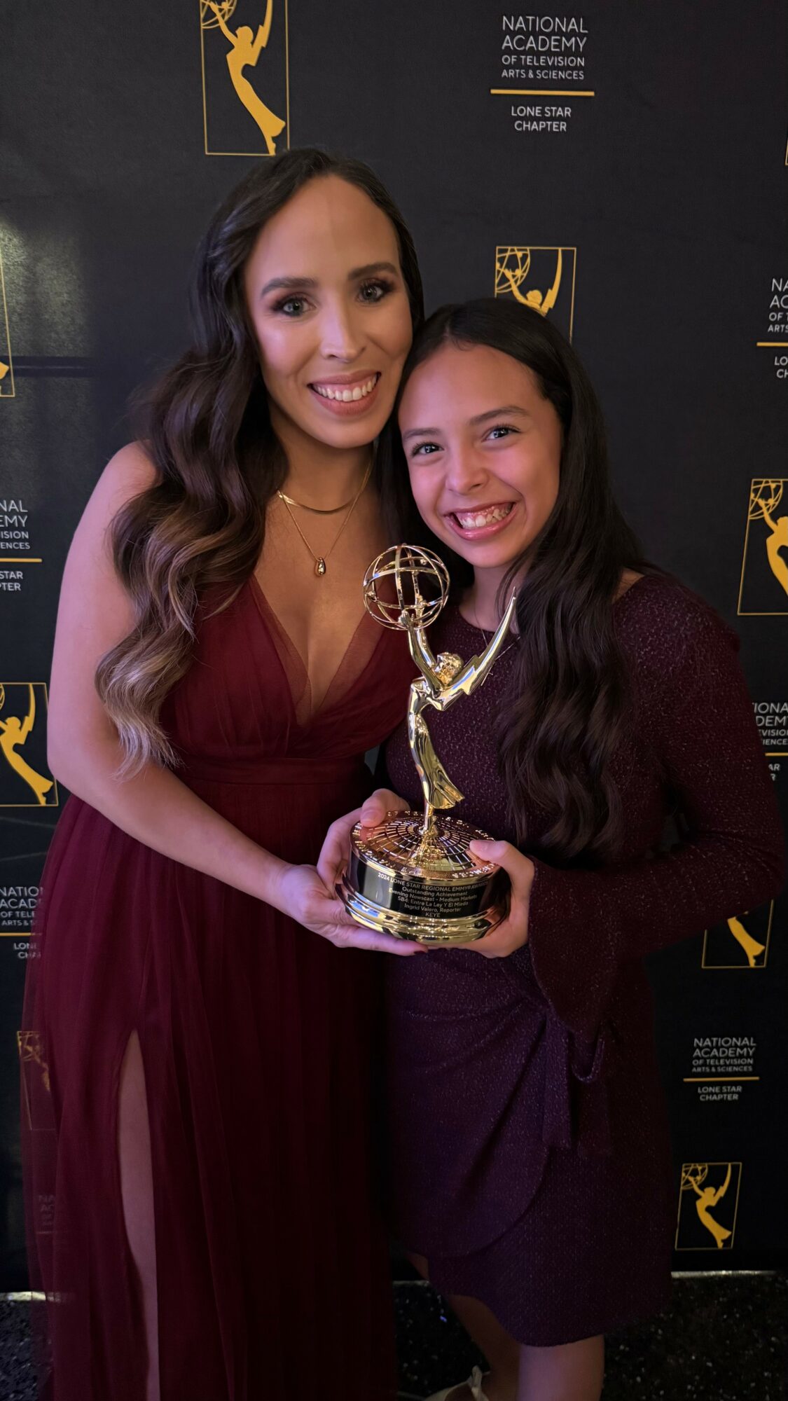 Two women smiling, one holding an Emmy award, standing in front of a black backdrop with gold Emmy logos.