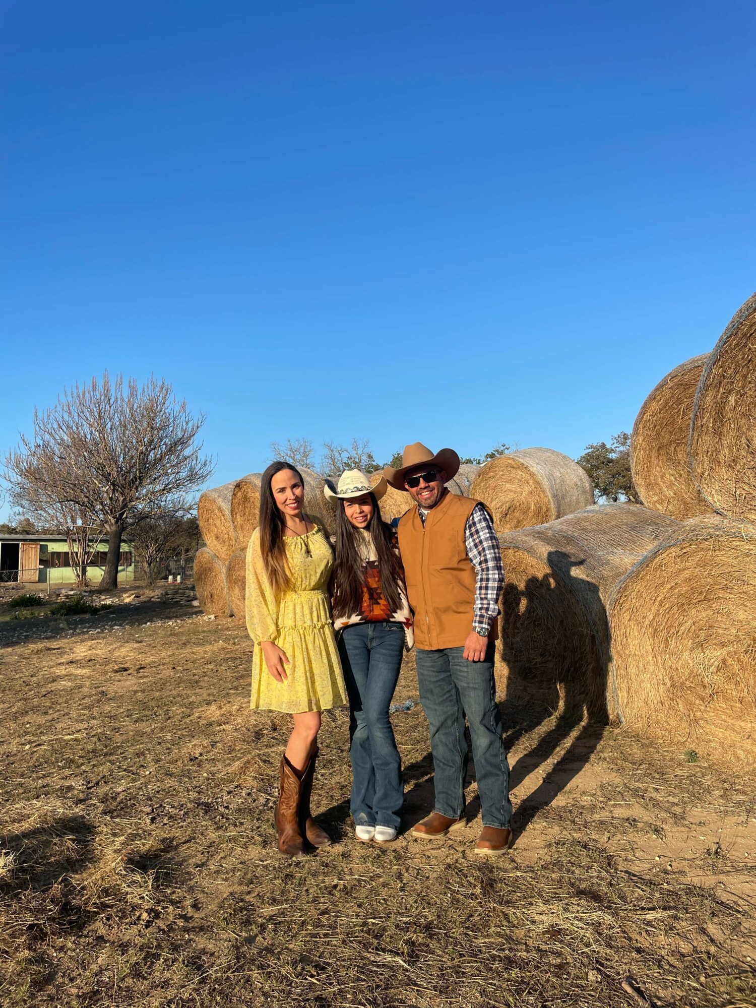 Three people standing outdoors near hay bales, with a clear blue sky and a leafless tree in the background.
