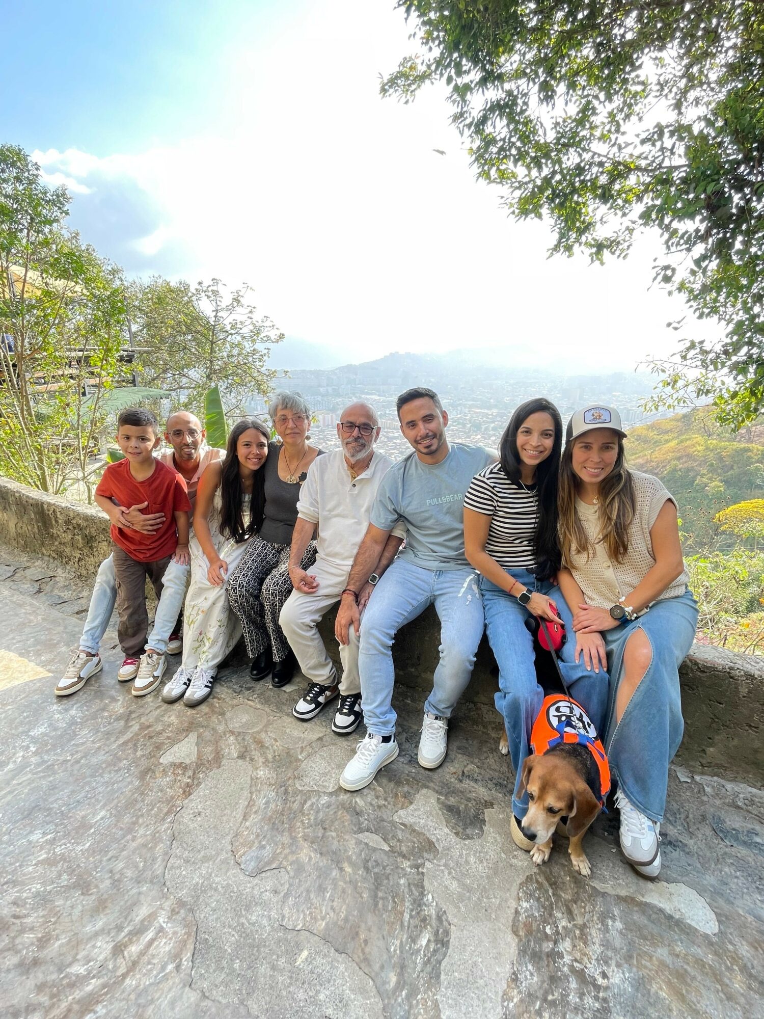 Group of seven people and a dog sitting on a rocky outdoor area with trees and sky in the background.