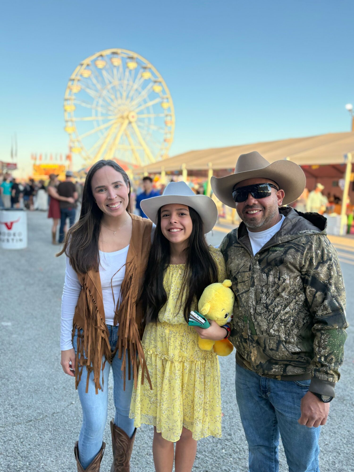 Three people smiling at a fairground with a Ferris wheel in the background, one holding a yellow plush toy, under a clear blue sky.