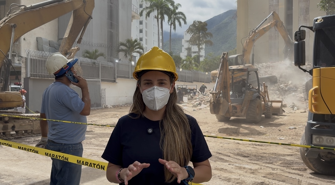 Woman wearing a yellow helmet and face mask at construction site with machinery and dust, person in background talking on phone.