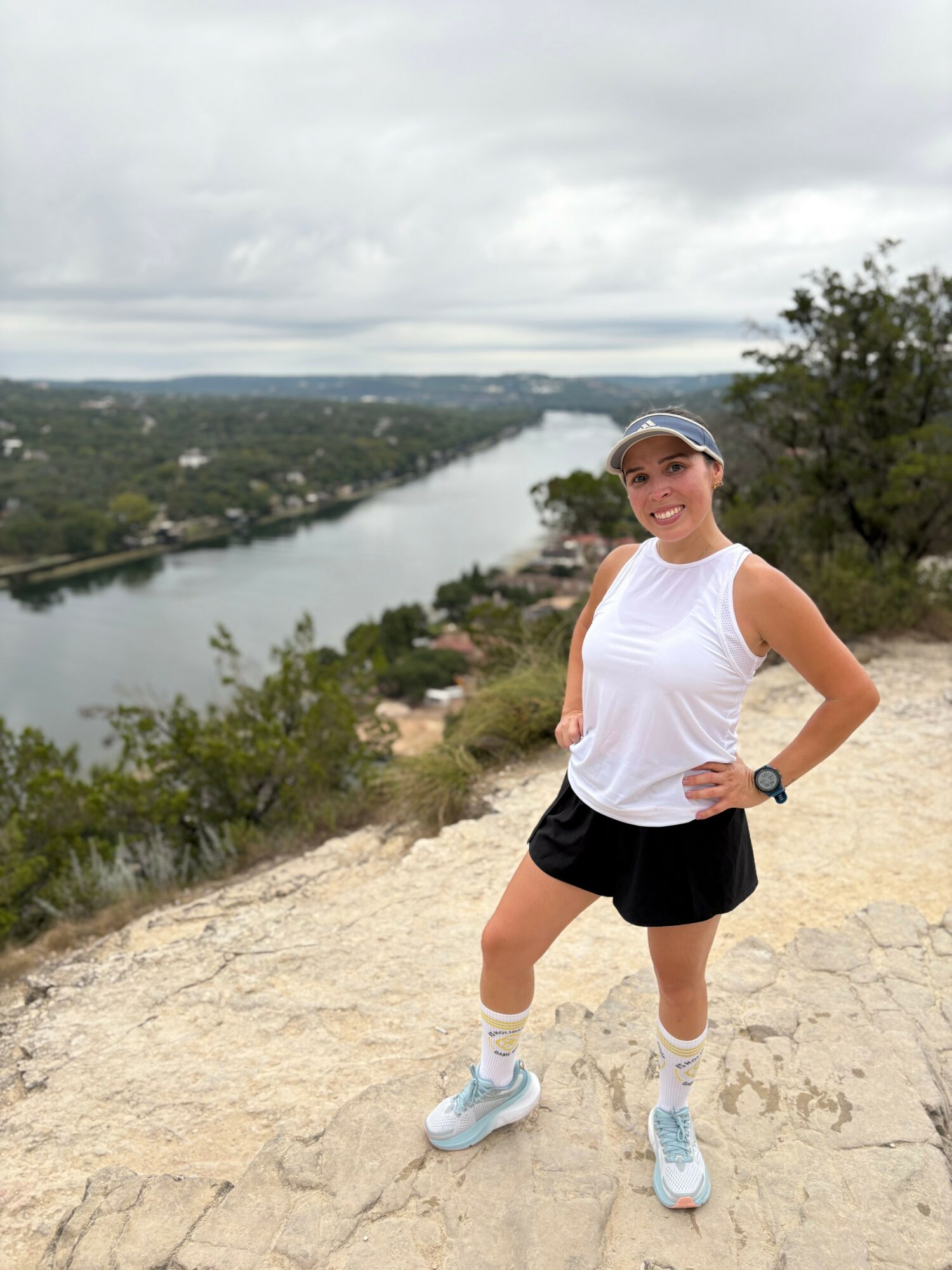 Smiling woman in athletic clothing standing on a hill with a river and landscape in the background.