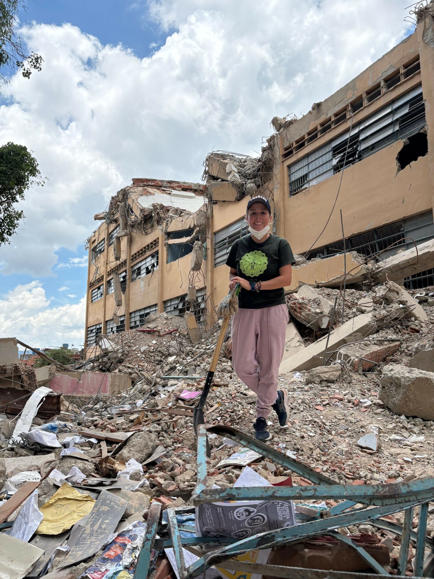 Woman standing among rubble and debris in a damaged urban area, with buildings and a cloudy sky in the background.