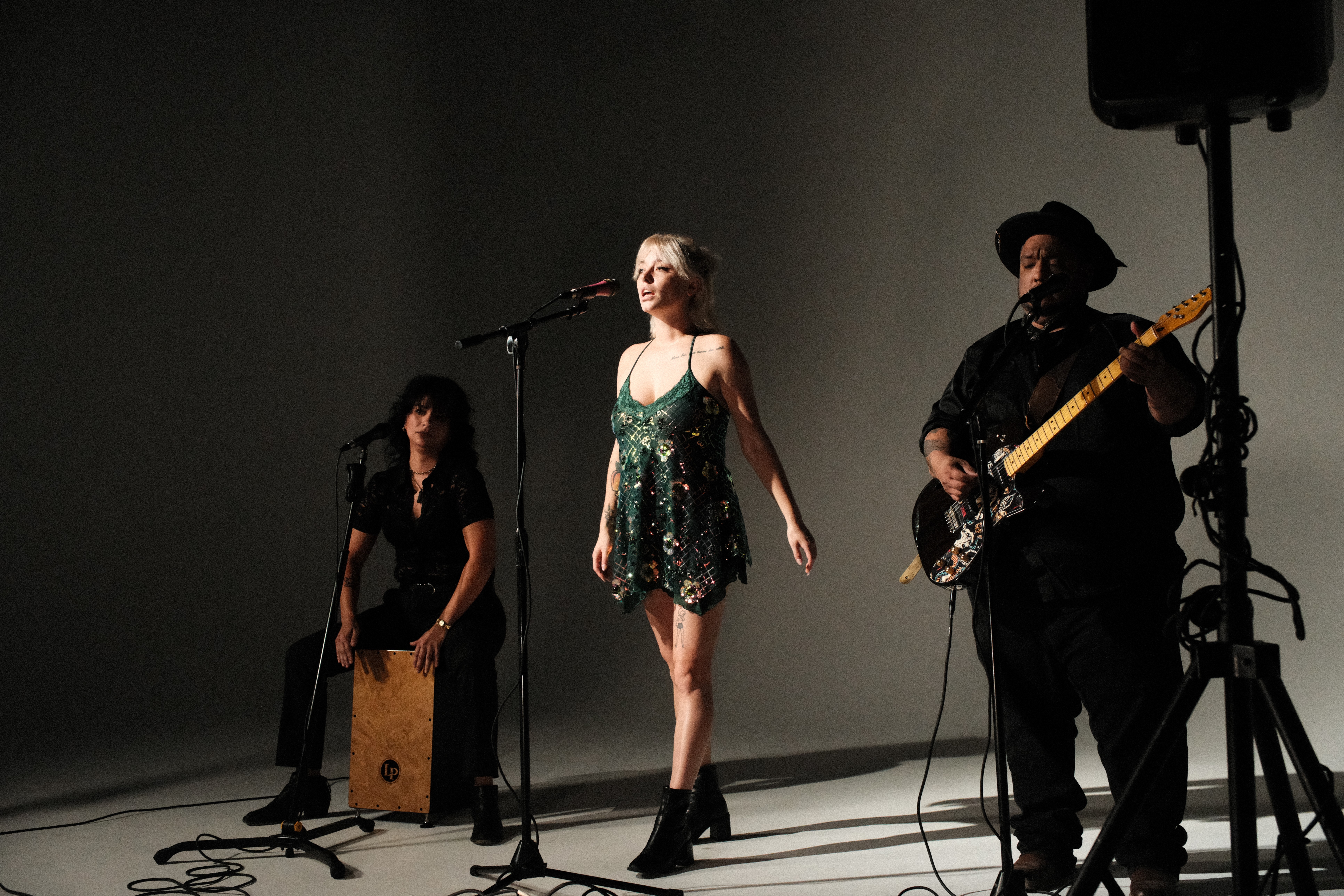 Three performers on stage: a woman singing at microphone, a man playing guitar, and a person sitting with a percussion instrument.