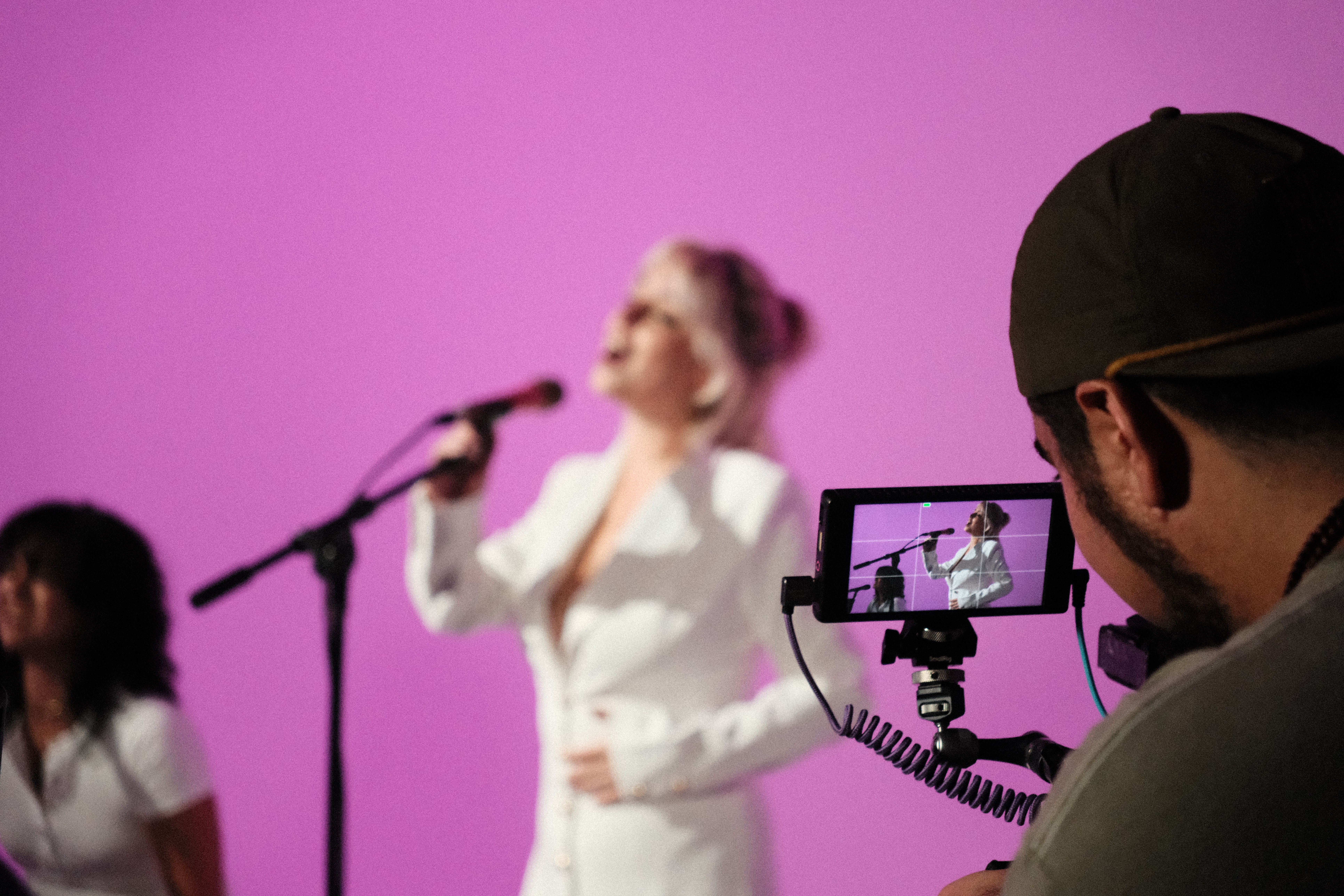 Person filming a woman singing on stage with a pink background, wearing a black cap and glasses, close-up view.