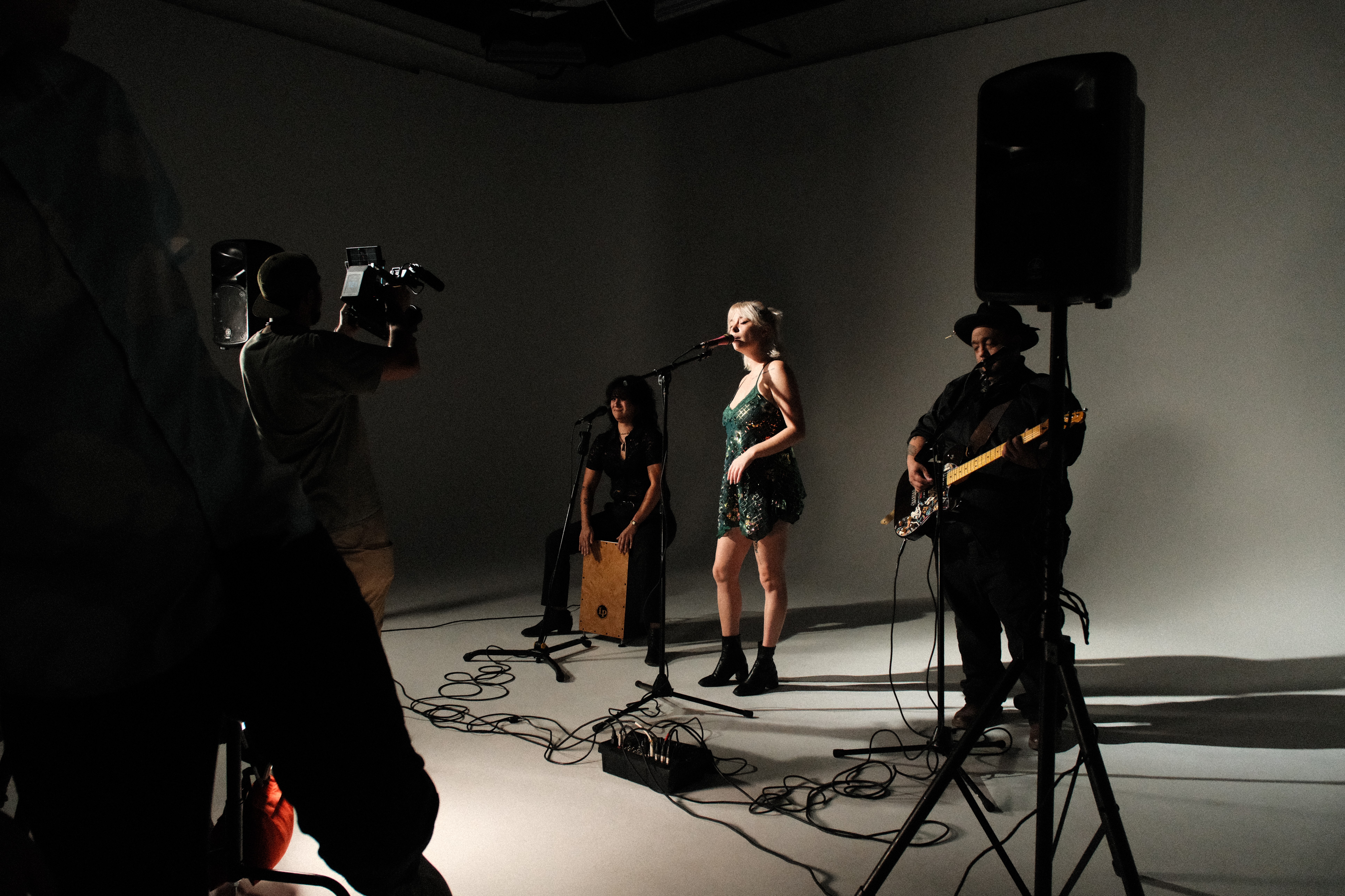 A woman performs on stage with a band, including a guitarist and percussionist, in a minimal studio setting.