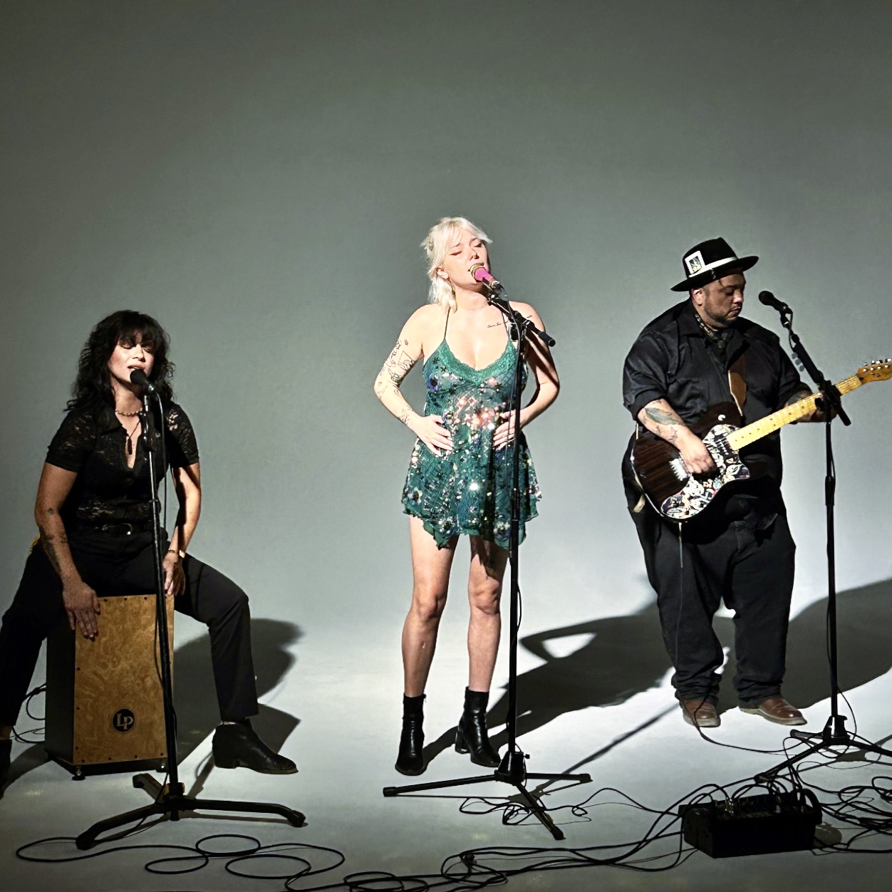 Three musicians perform on stage with microphones, a cajón, and an electric guitar, against a plain background.