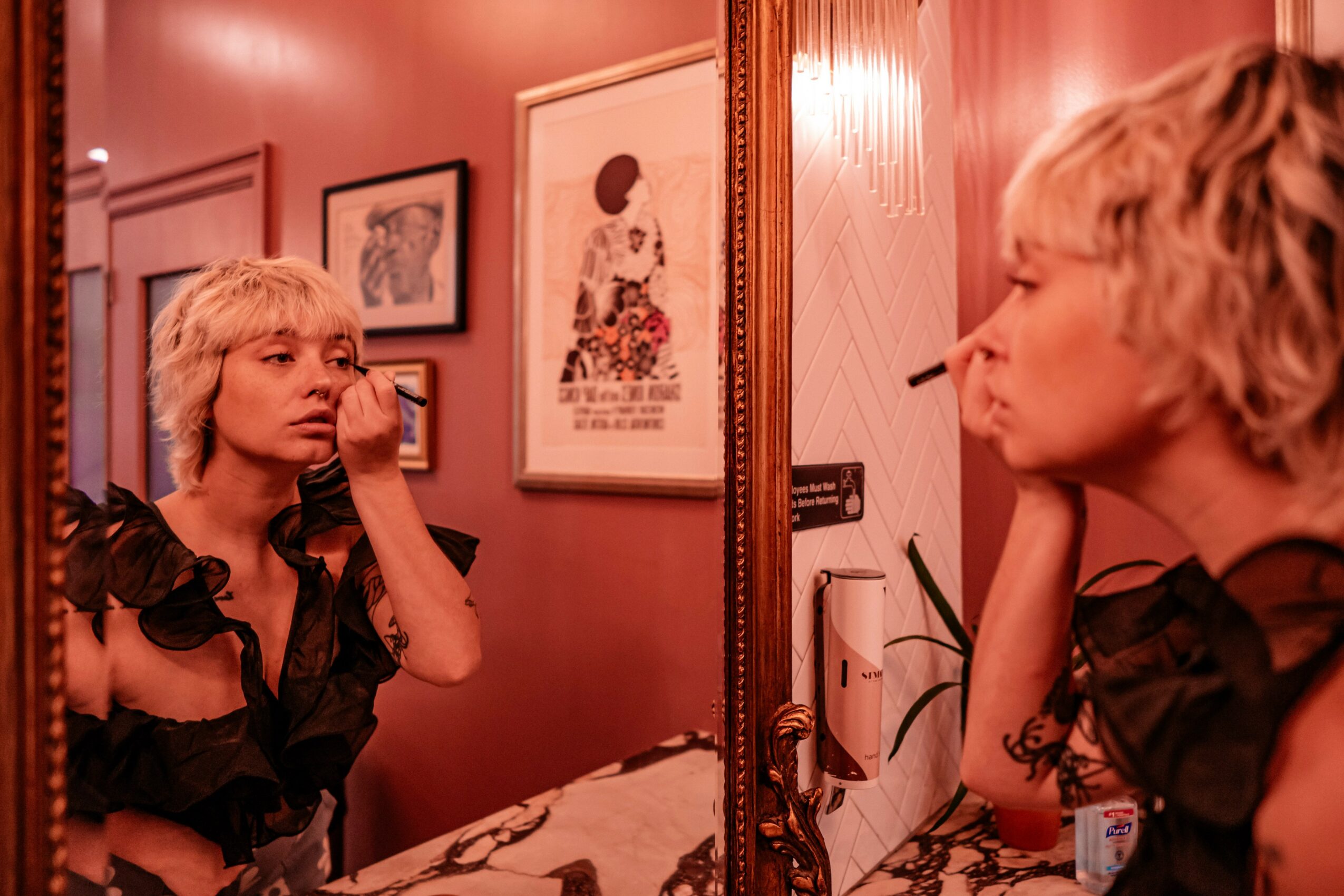 Woman with short blonde hair applying makeup in front of a mirror, with framed pictures on the wall behind her.
