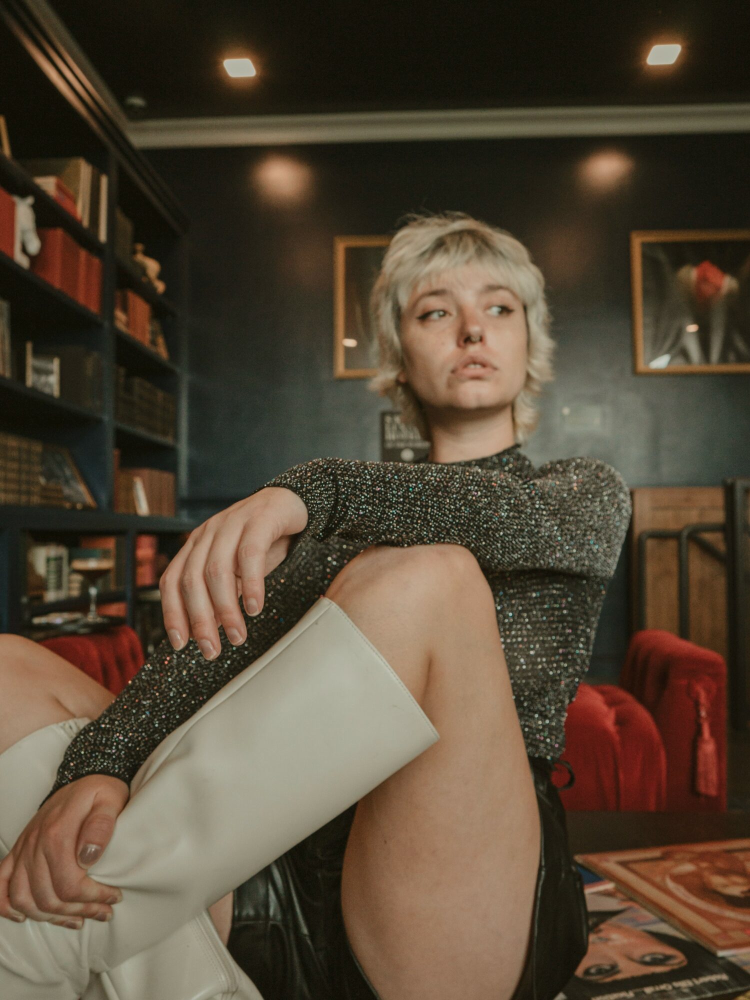 Young woman with short blonde hair sitting in a dimly lit room, holding a white bag, with bookshelves and framed pictures behind her.