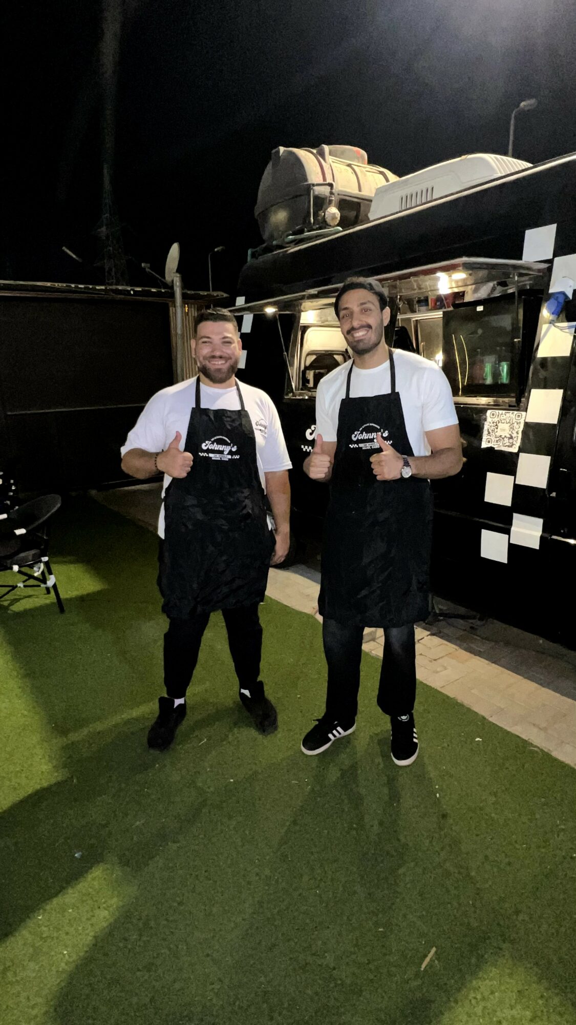 Two men stand outdoors in front of a food truck, smiling and wearing aprons, with a dark background and artificial grass.
