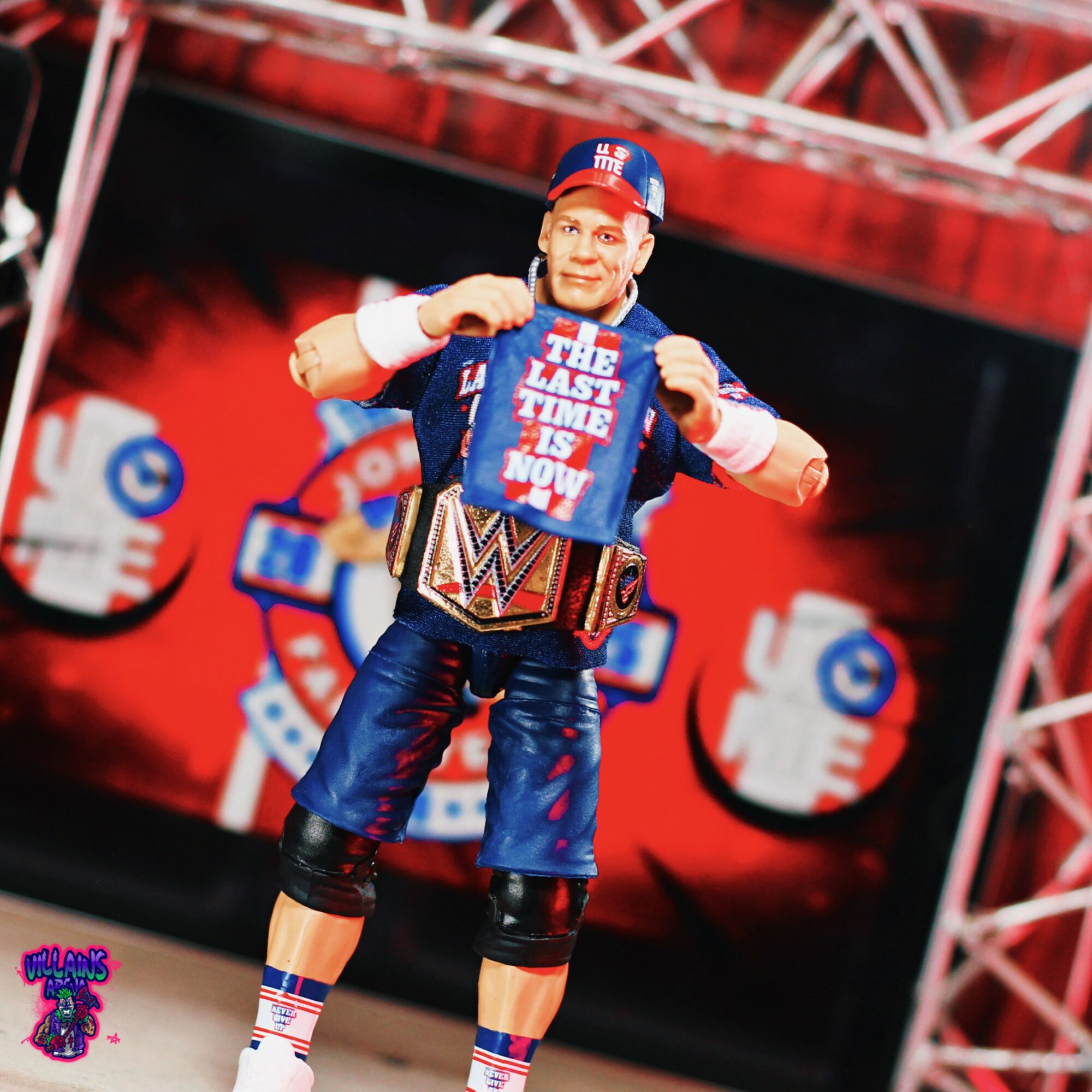 Person dressed as a wrestler with a championship belt, holding a sign, on a stage with red and black background.