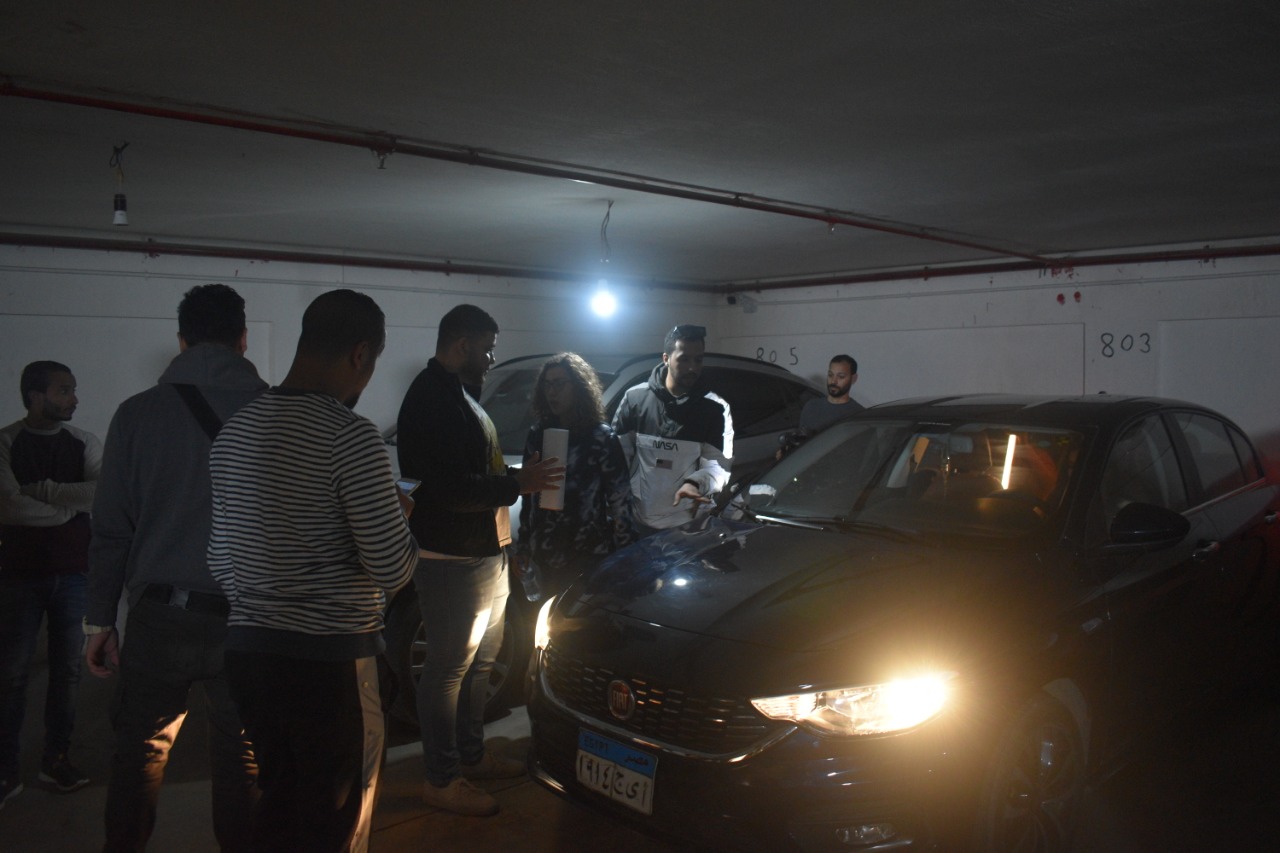 Group of people gathered around a car in a dimly lit underground parking garage, some inspecting the vehicle, with bright headlights on.