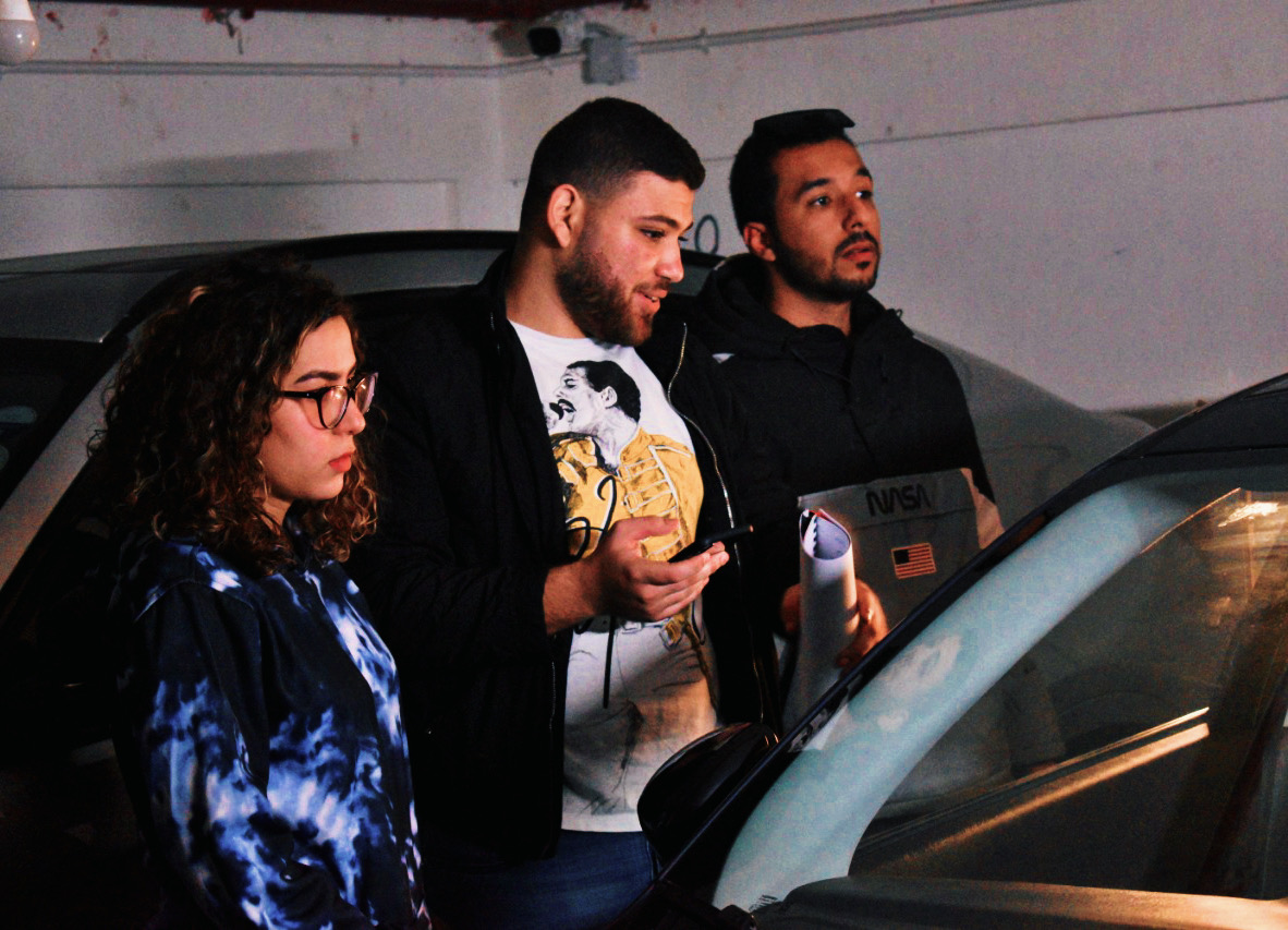 Three people standing outdoors near a vehicle, looking at a device or screen, with a wall in the background.