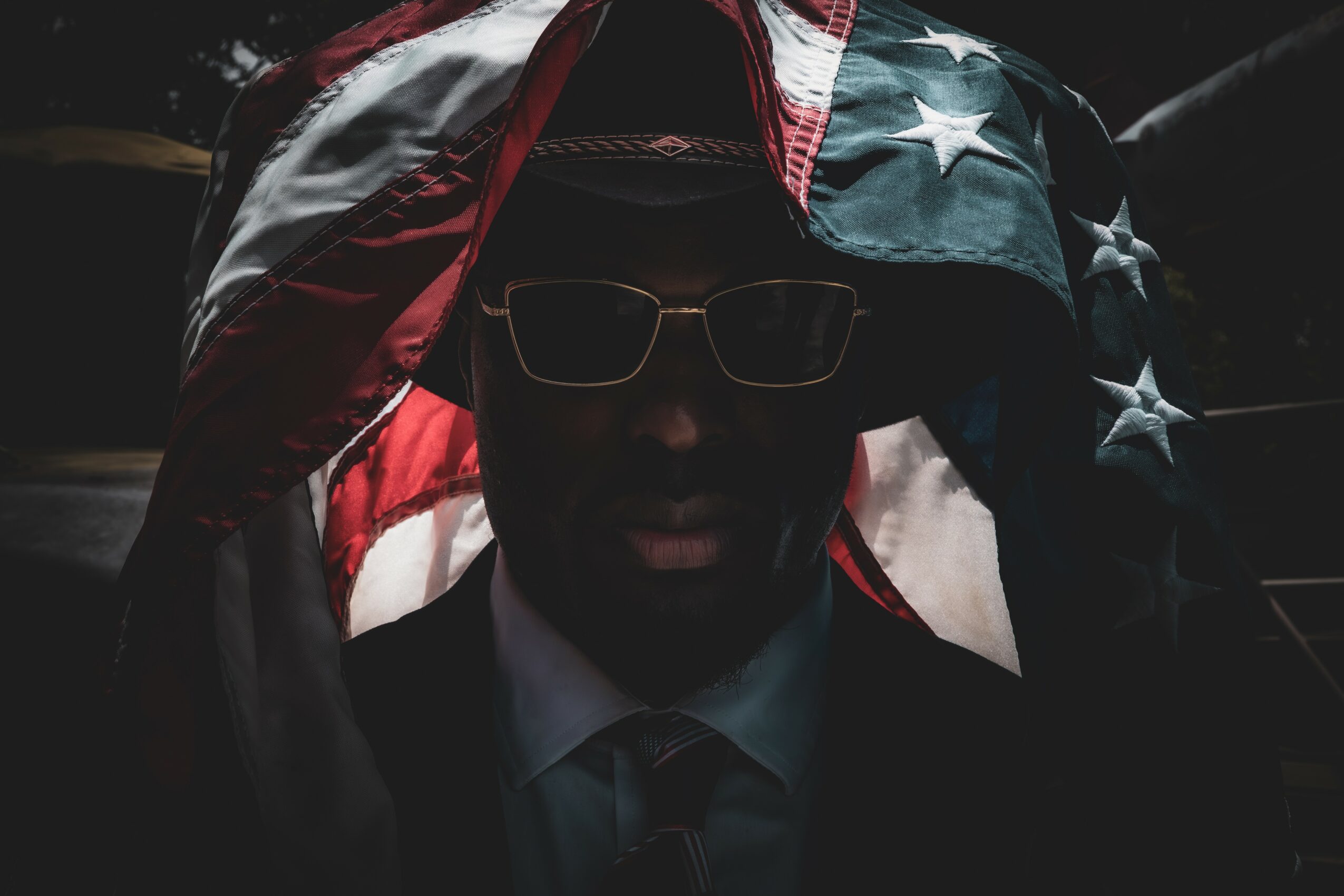Person wearing glasses with an American flag draped over their head, dark background, face partially visible.