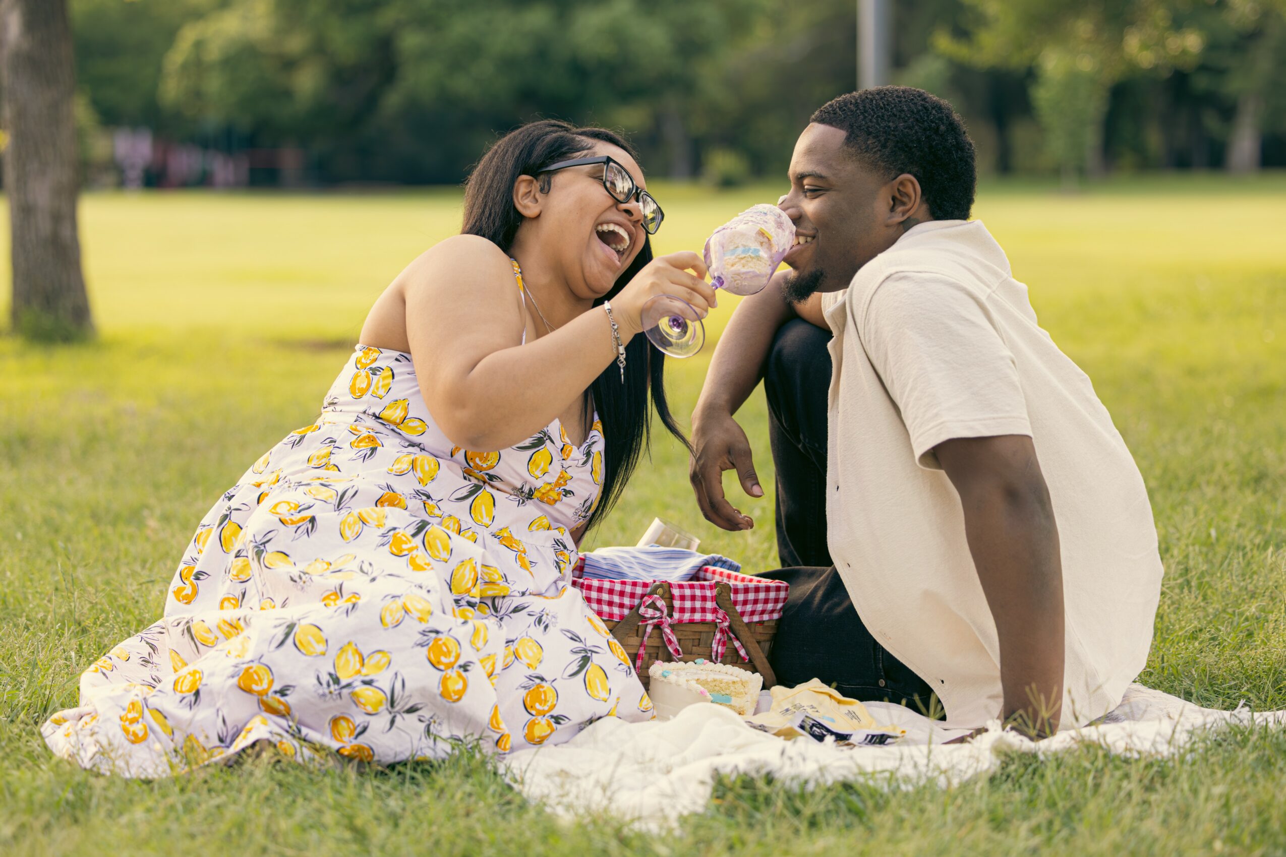 Two people sitting on a blanket in a park, sharing ice cream and smiling, with trees and grass in the background.